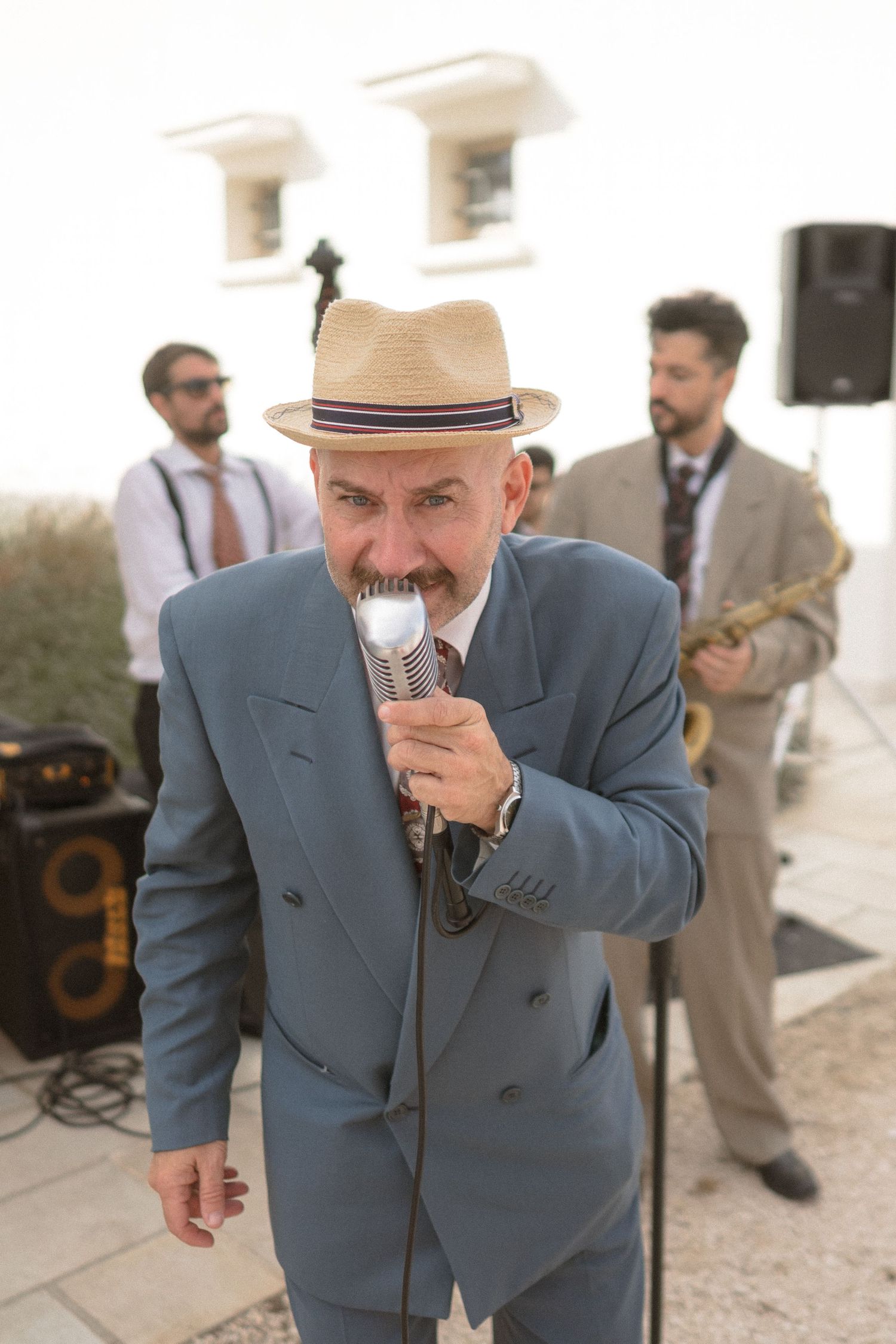 Man in vintage-style blue suit and fedora hat takes a drink at an outdoor event.