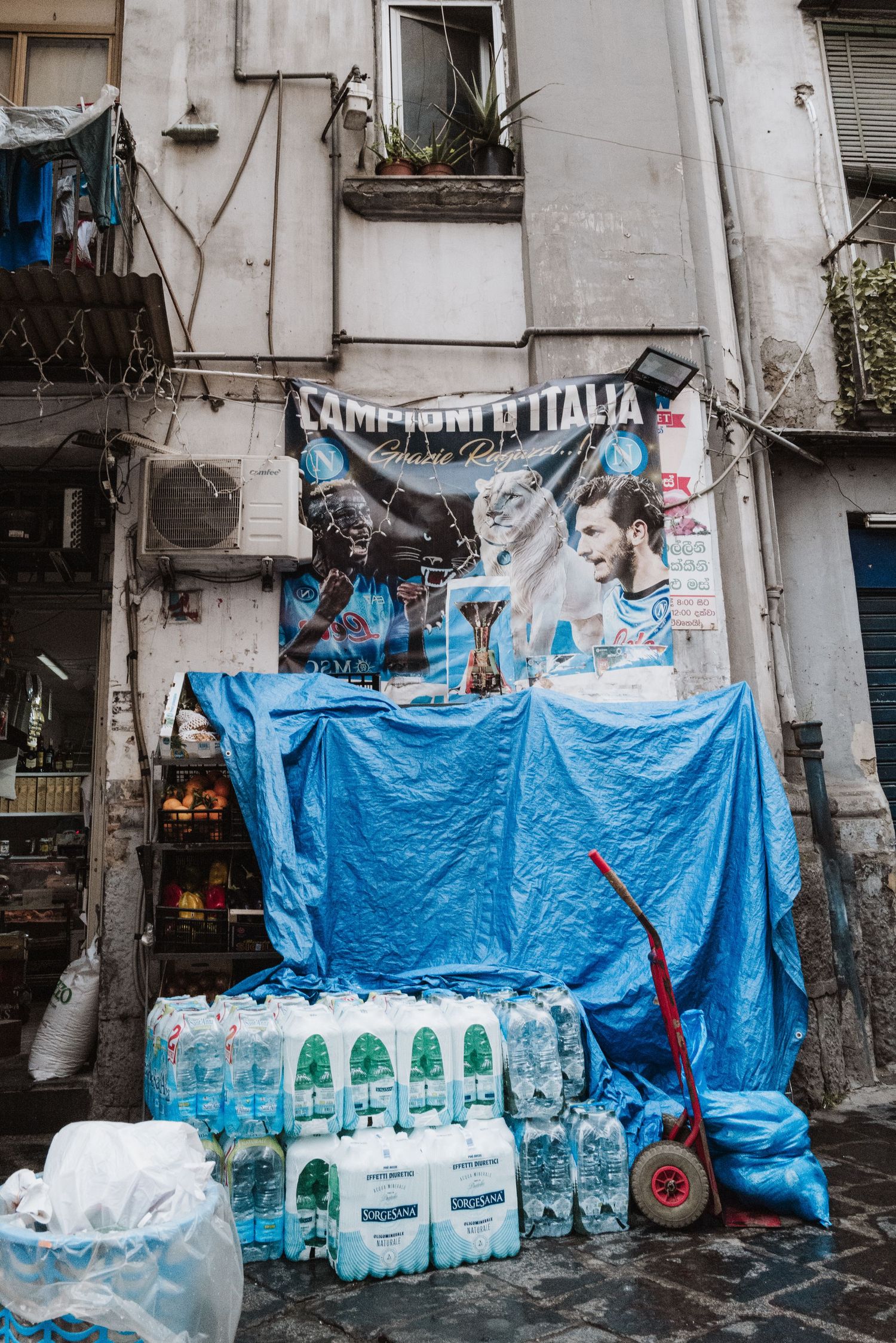 A display of water bottles and blue tarp against a weathered building wall with graffiti in Naples.