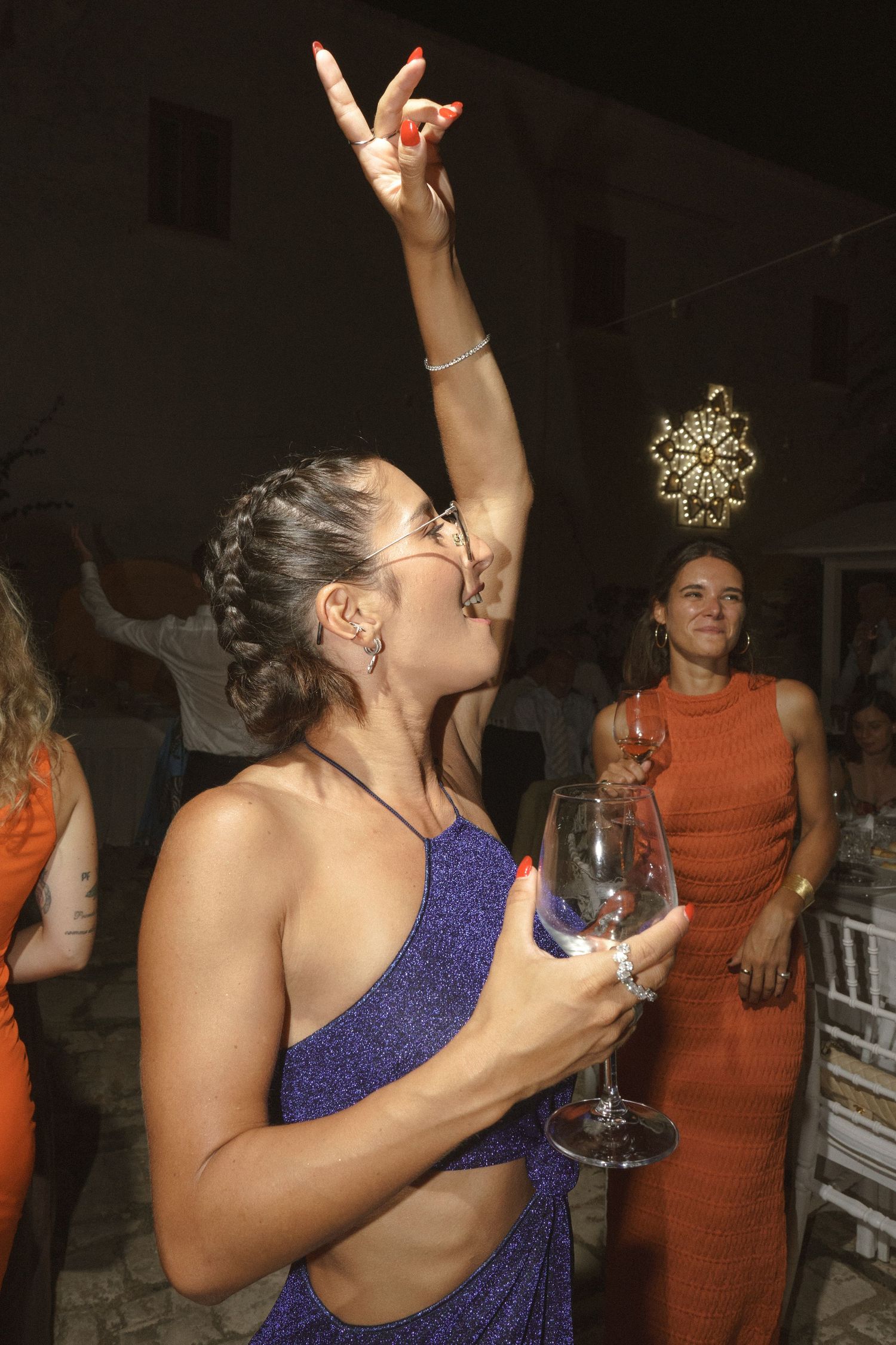 Person in a sparkly purple dress dancing with a wine glass at a party while someone in orange looks on.