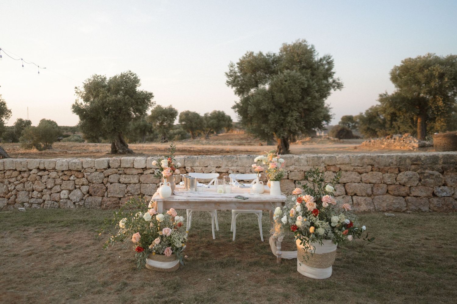 Elegant outdoor table setting with floral arrangements against a rustic stone wall at sunset.