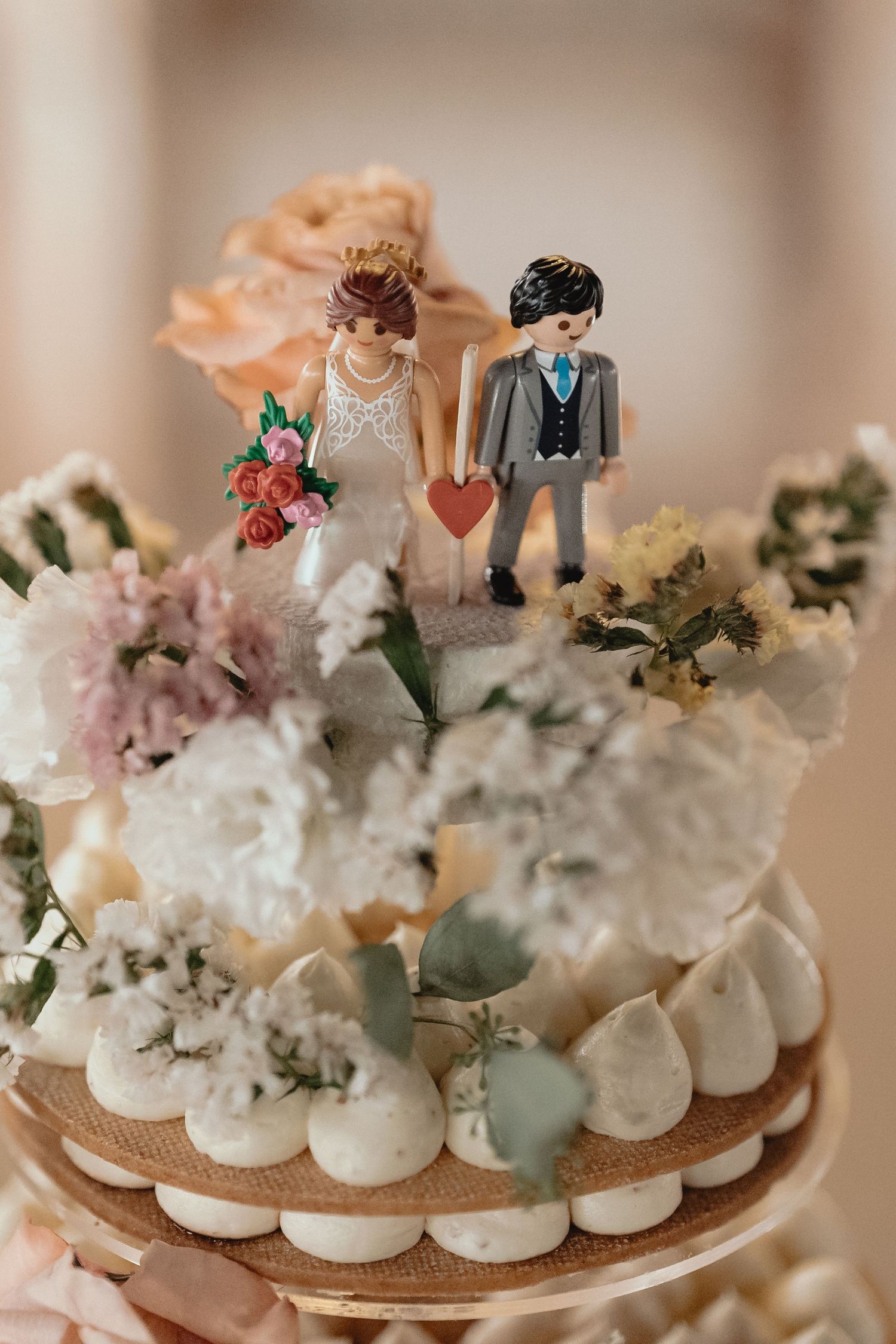 Delicate meringue desserts decorated with pastel flowers and miniature wedding cake toppers on a display stand.