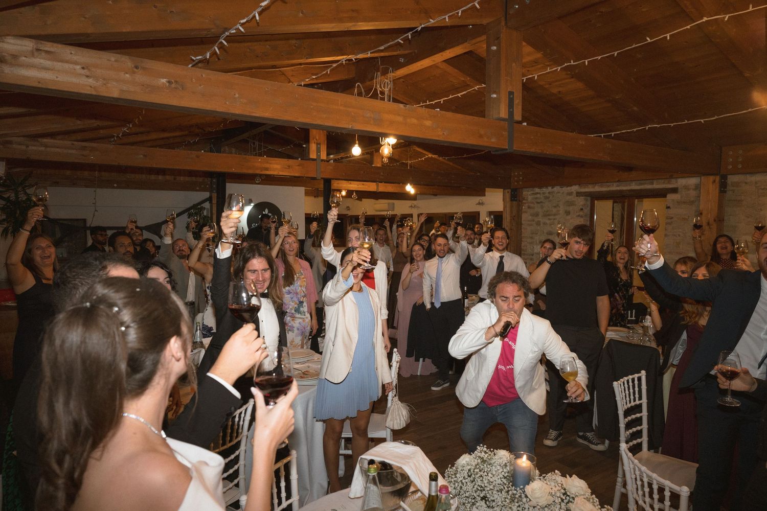 Wedding reception guests dancing and celebrating on a rustic wooden dance floor under warm lighting.