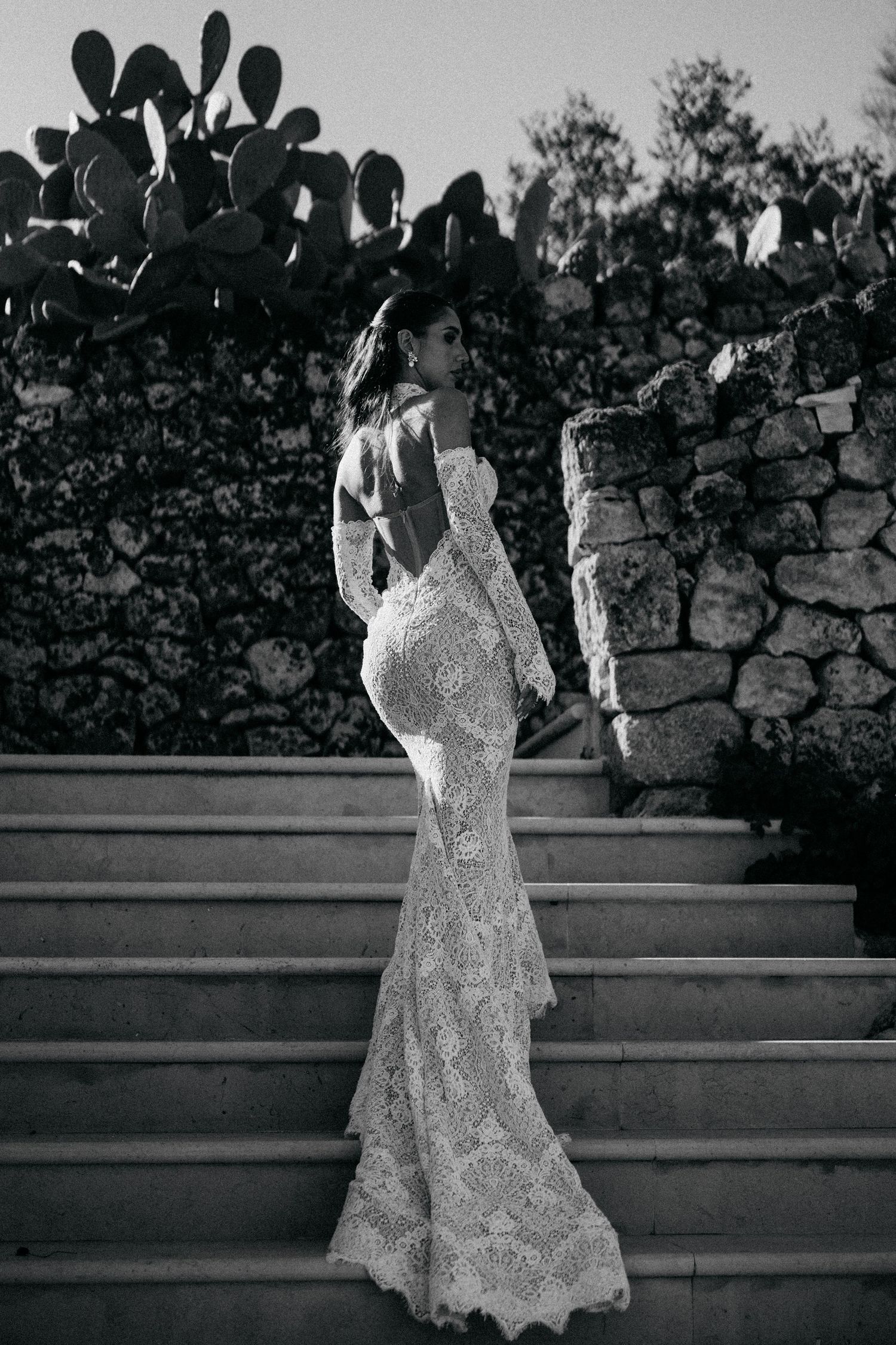 Black and white image of a bride in a backless lace mermaid gown climbing stone steps beside a rustic wall.