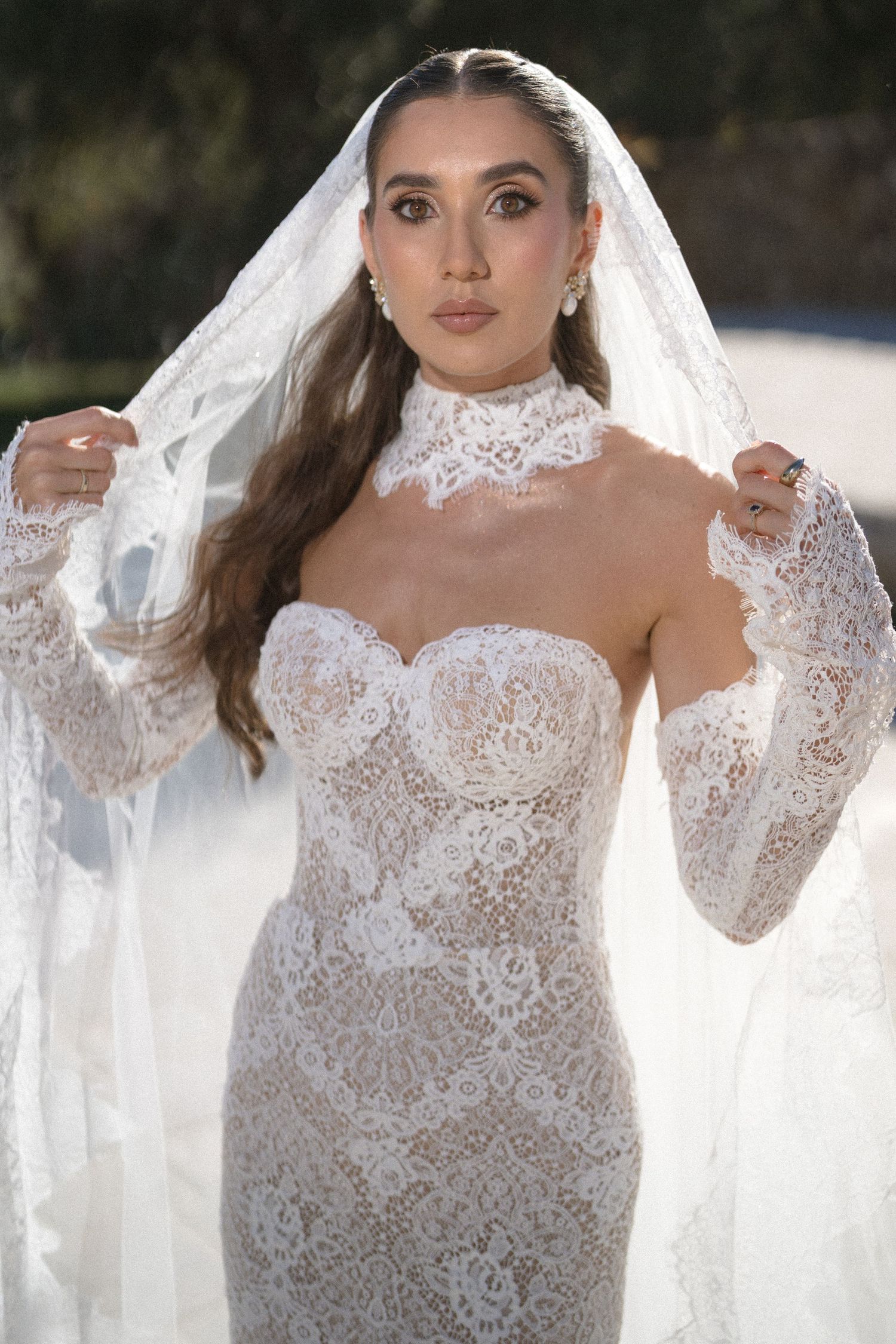 Smiling bride holding her sheer lace-edged veil while wearing a strapless lace wedding dress with a high lace choker collar.