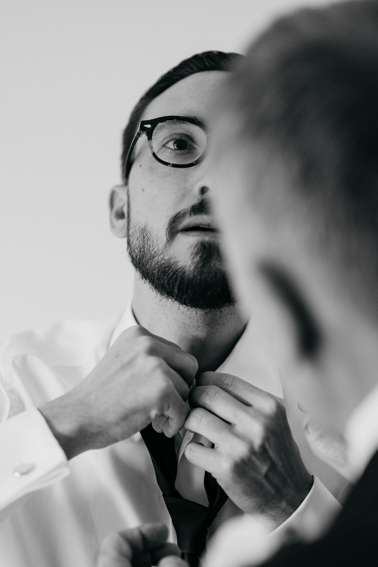 A groom adjusts his tie while looking in the mirror, wearing glasses and a white dress shirt, in a black and white photo.