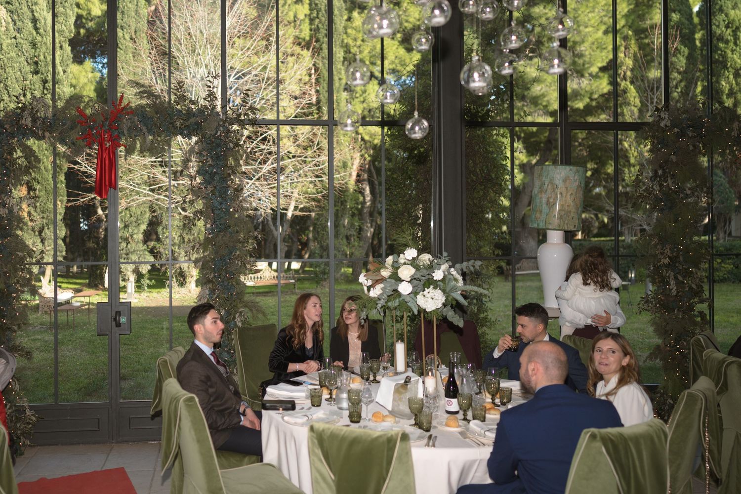 Wedding reception guests dining in elegant glass greenhouse venue with green floral centerpieces.