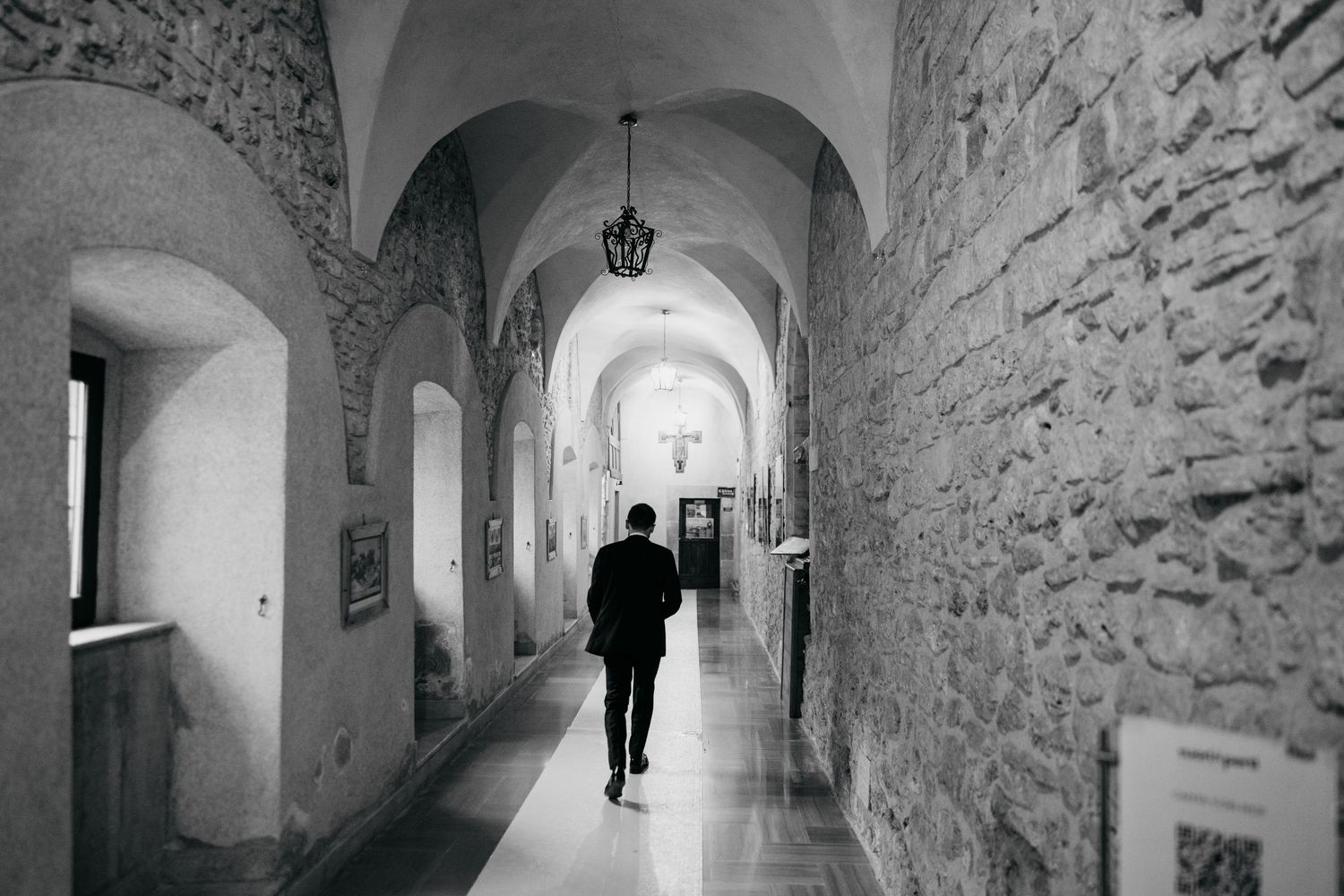 Man walks alone through arched stone corridor in black and white wedding photo.