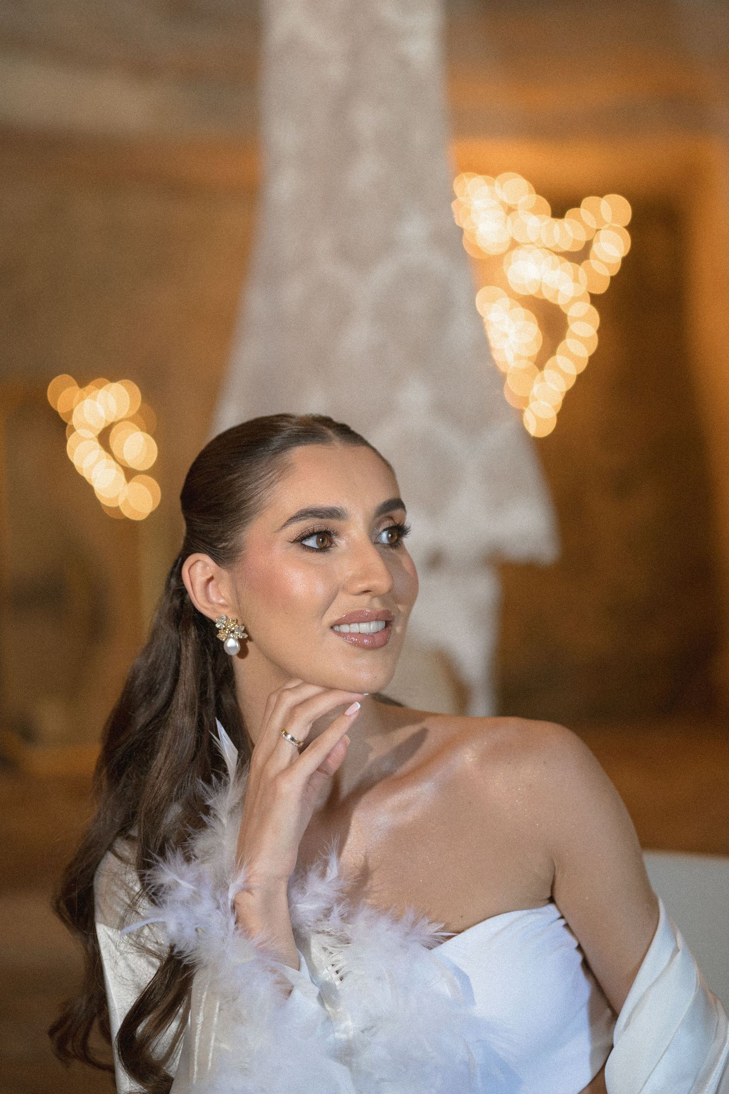 Smiling bride with a sleek ponytail, pearl earrings and a white feathered wrap poses beside warm heart-shaped lights.
