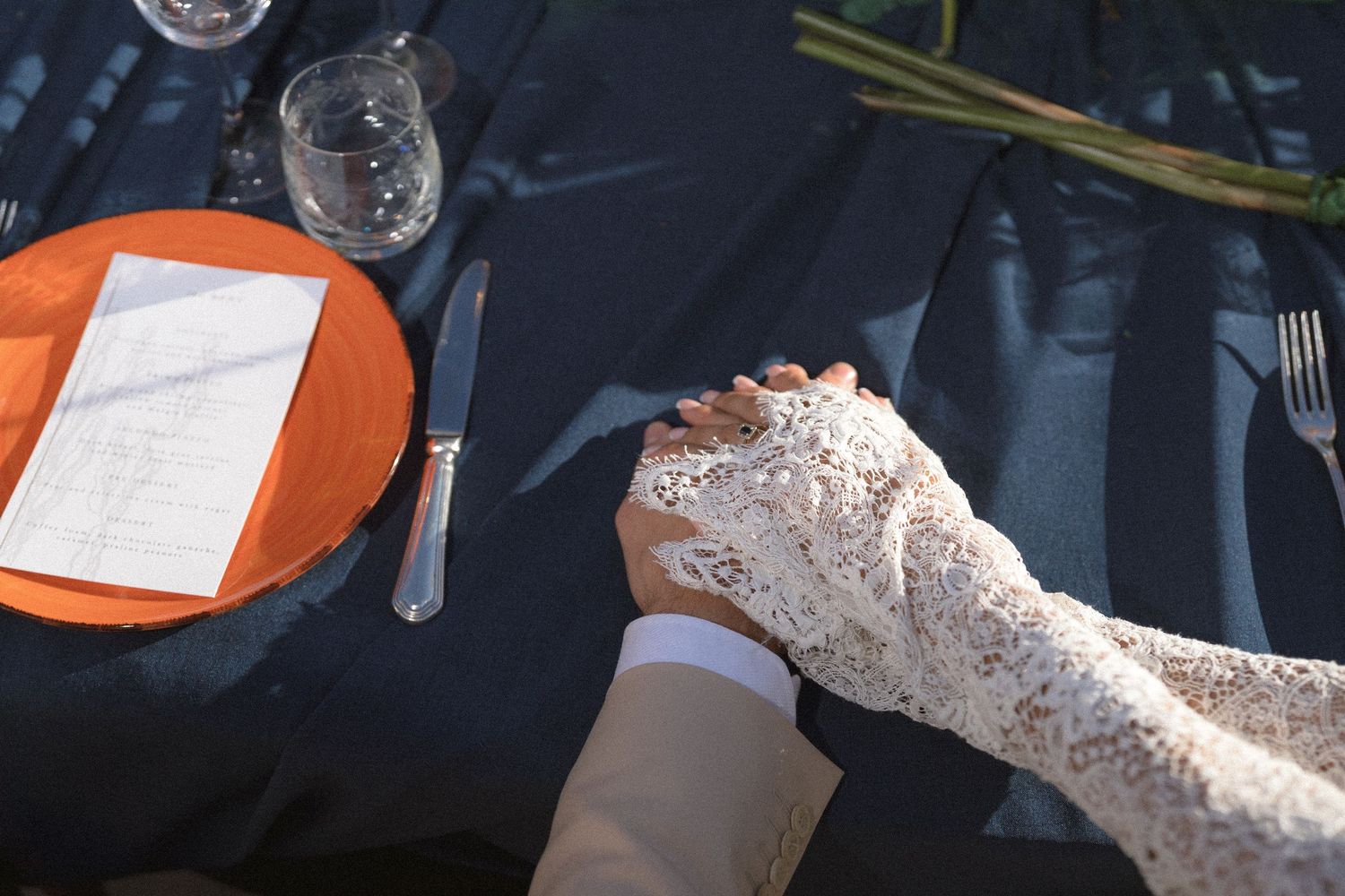 Bride in a delicate lace sleeve holds the groom's hand on a dark tablecloth beside a place setting and menu card.