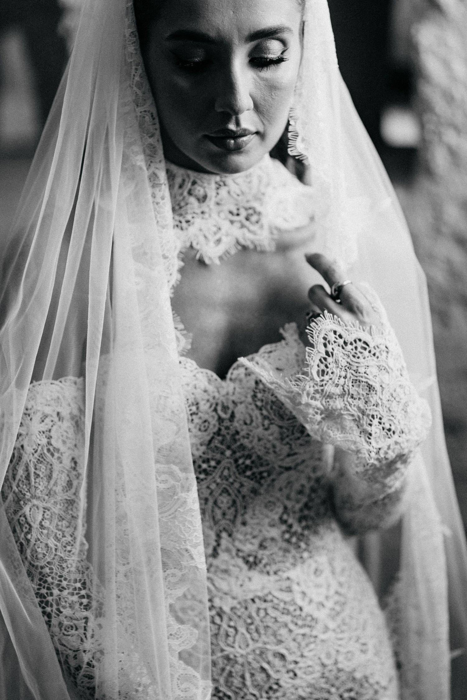 Black and white portrait of a bride in a lace gown and sheer veil, looking down with her hand resting near her collar.