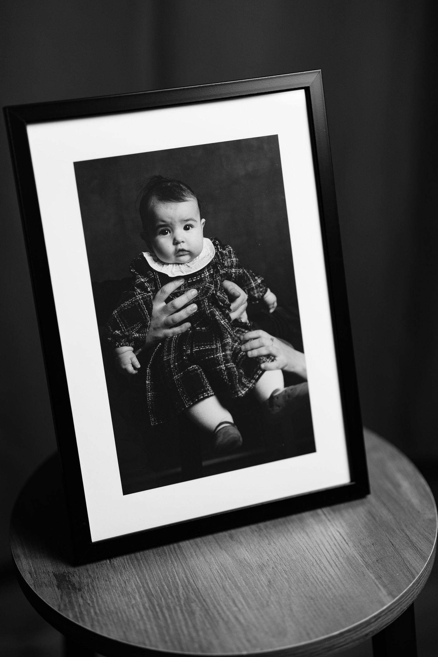 Black and white framed photo of a baby in plaid outfit displayed on a wooden table against dark background.