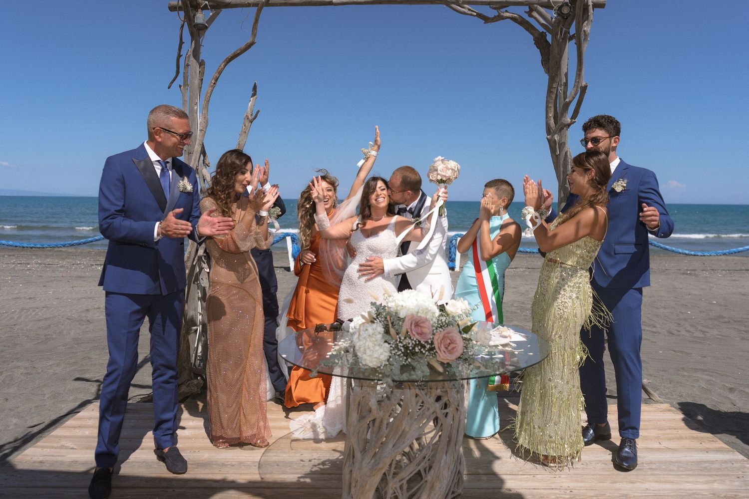 Wedding celebration on a beach with guests cheering under an ornate arch.