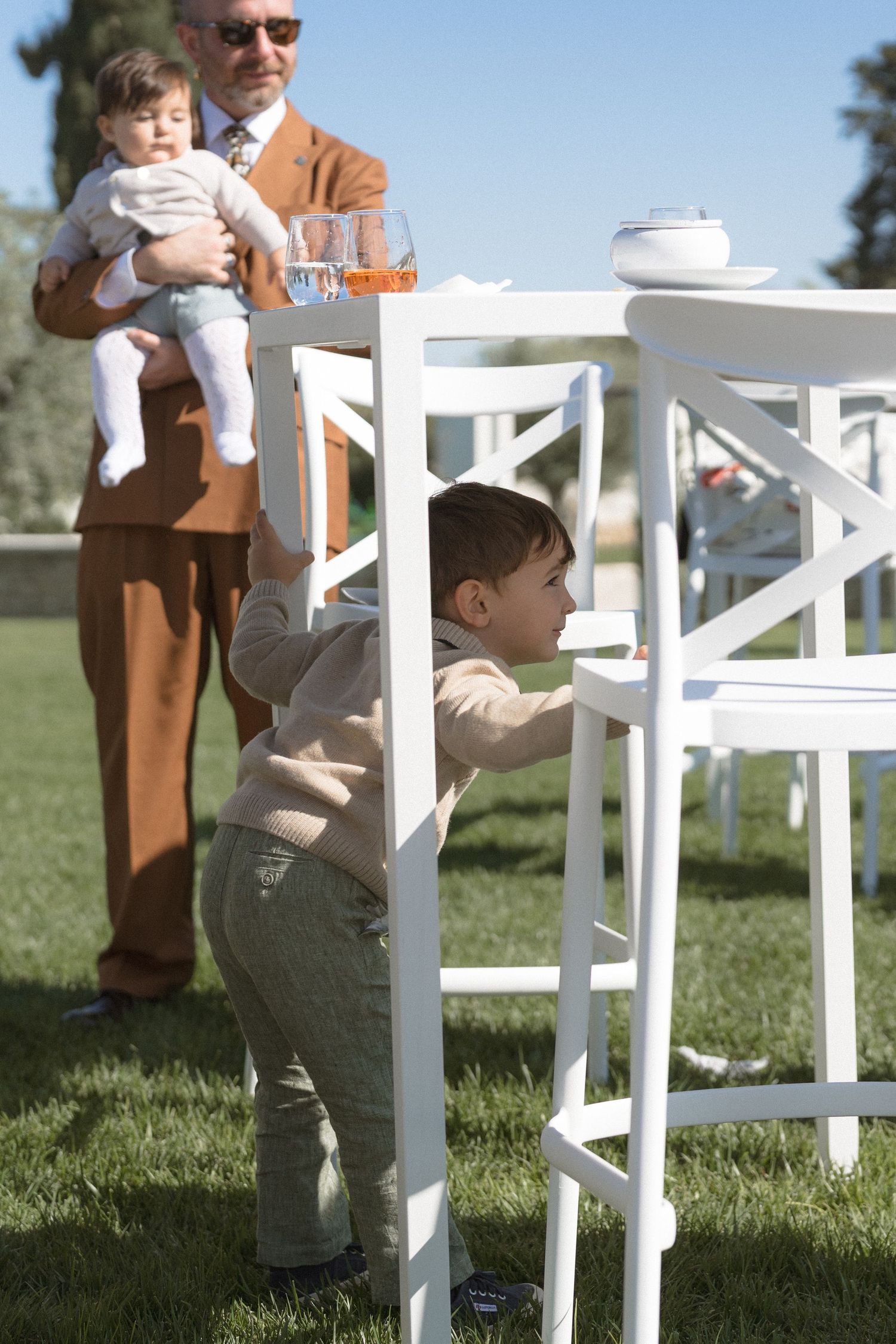 A young boy stands by a white outdoor chair while an older man holds a baby at a sunny garden event.