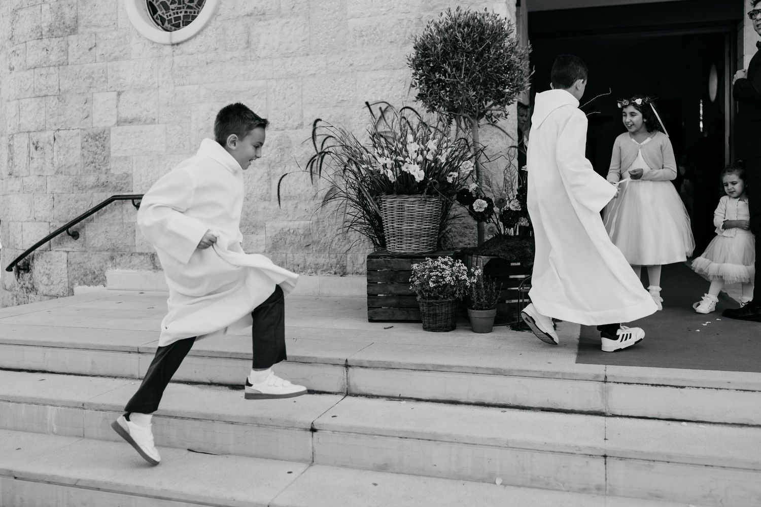 Children in white communion attire playfully running up stone church steps in a candid black and white photo.