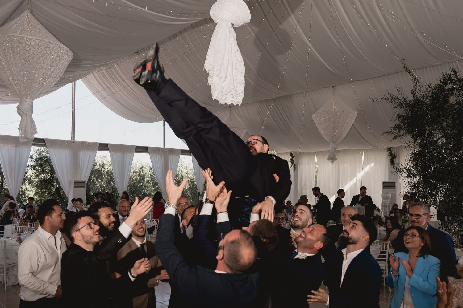 Wedding guests crowd together to lift someone up during a celebratory dance moment at an indoor reception.