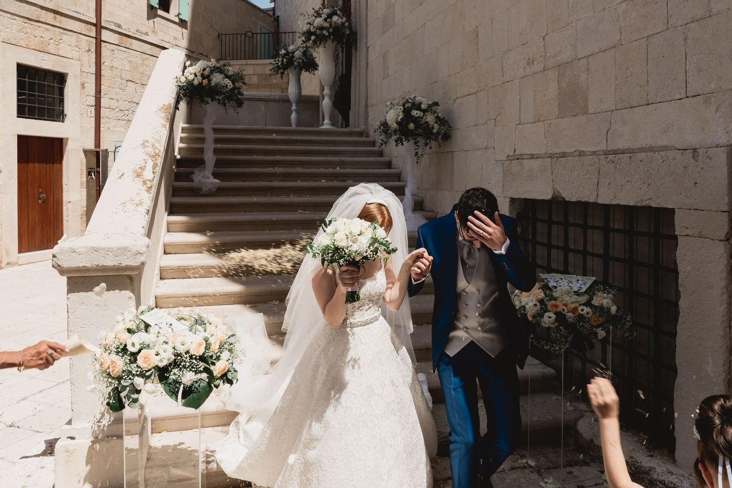 Couple shares playful moment on stone steps during outdoor wedding ceremony with white floral decorations.