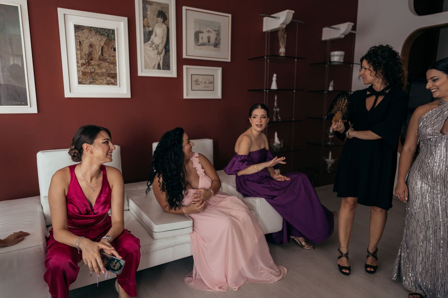 A group of women in formal dresses gathered in a room with red walls.
