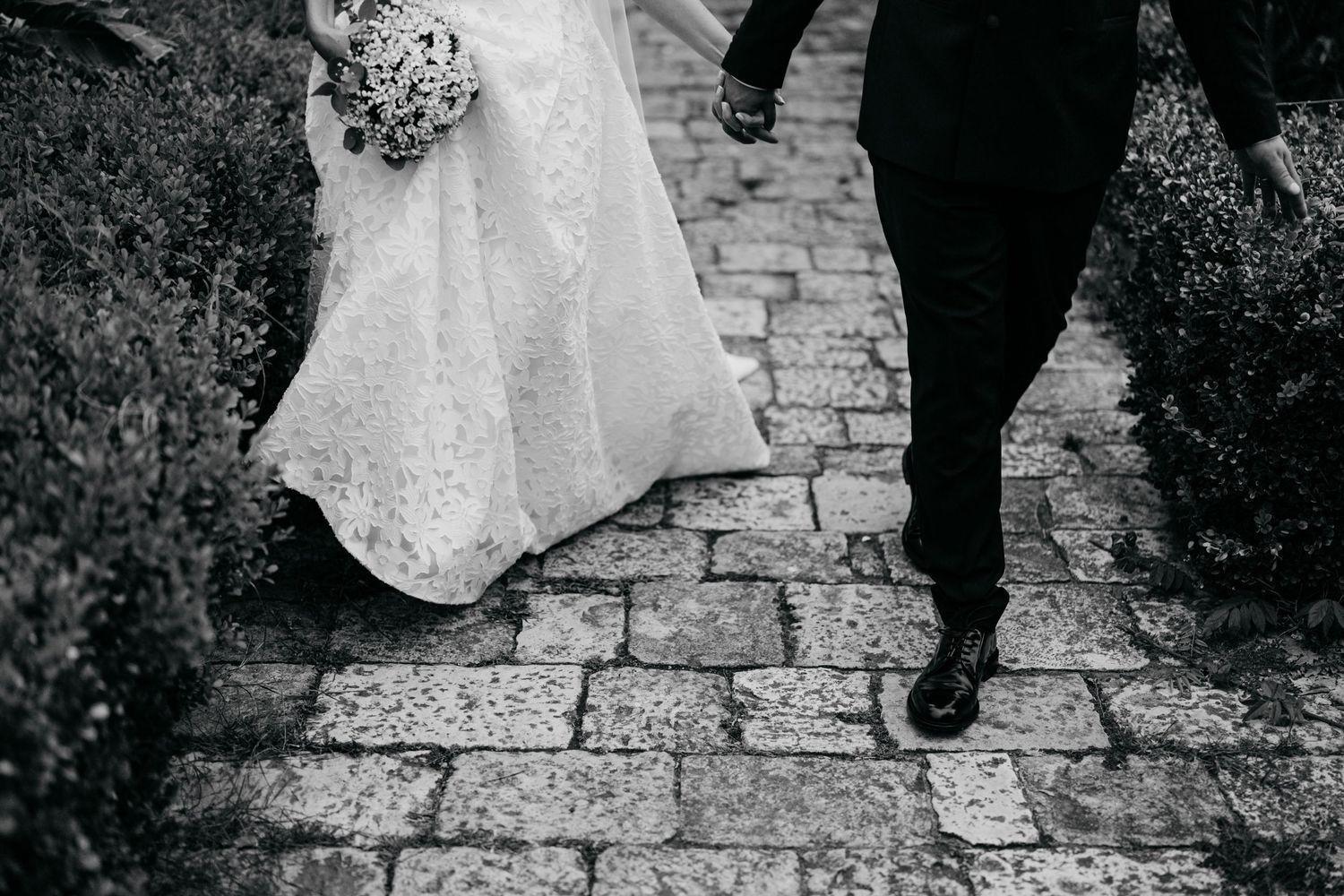 Black and white detail shot of wedding dress and formal shoes on stone pavement.