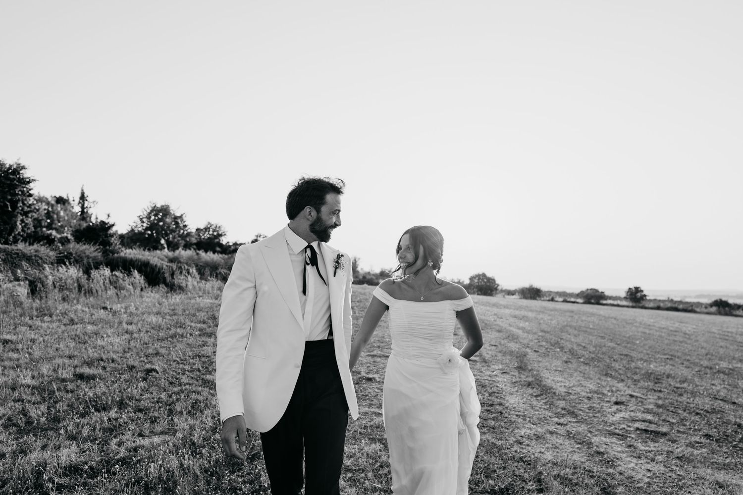 Black and white photo of couple walking hand in hand through an open field at sunset.
