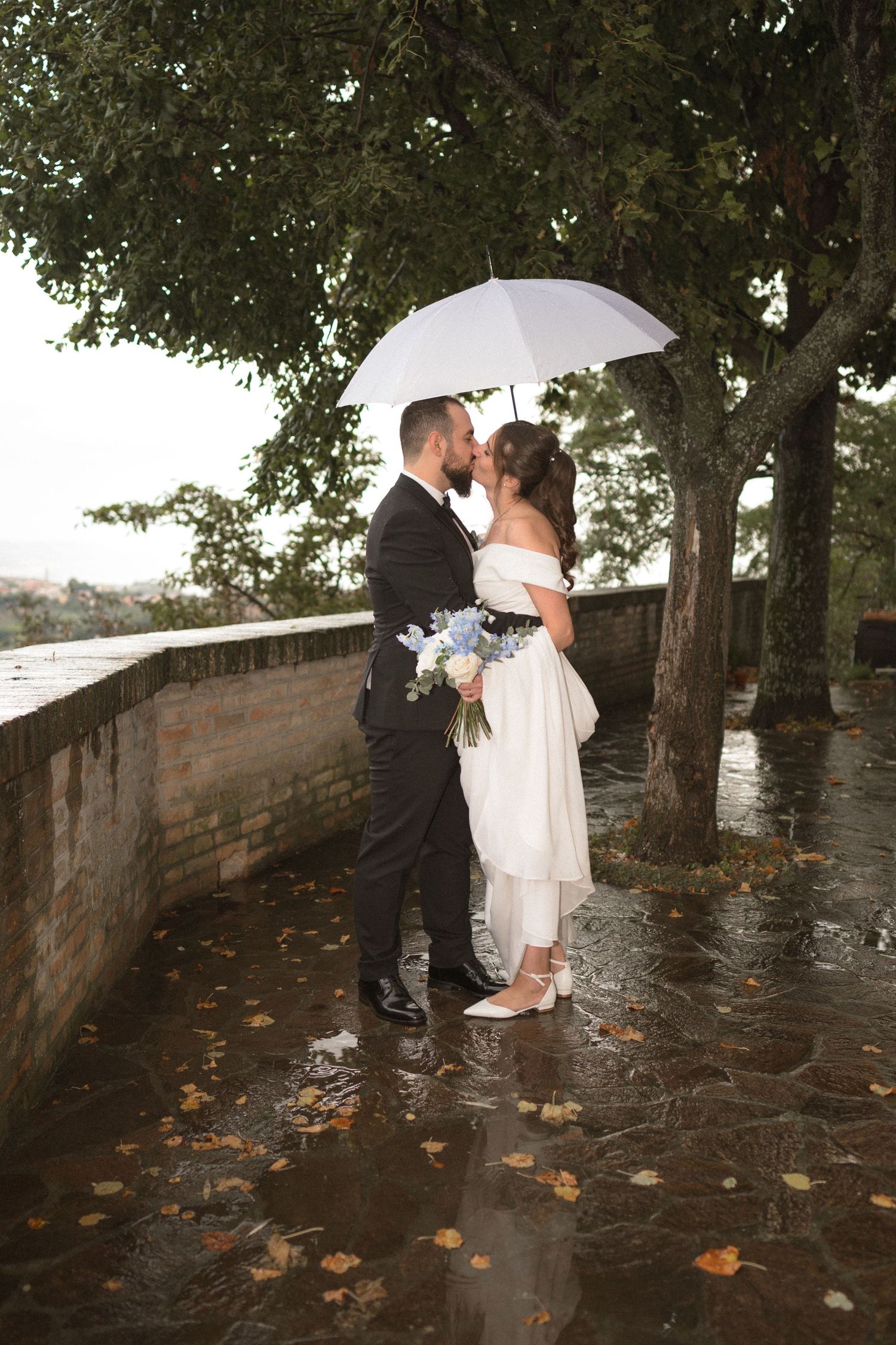 Newlywed couple shares intimate moment under trees in the rain with white umbrella during outdoor wedding photoshoot.
