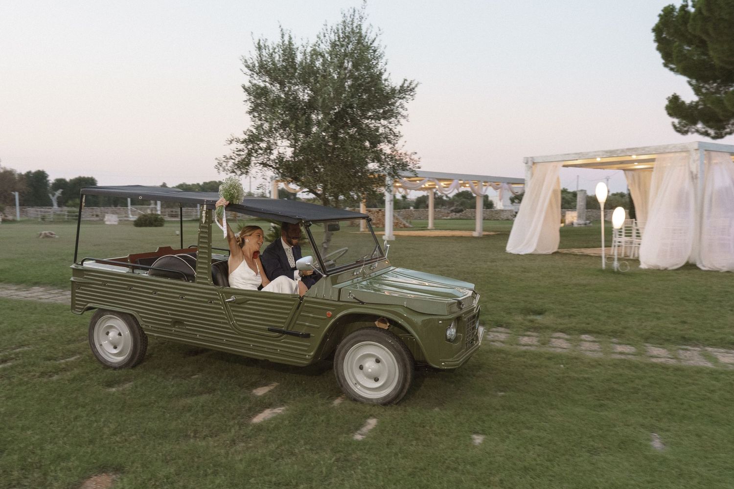 A vintage military-style jeep drives across a grassy field at a wedding venue.