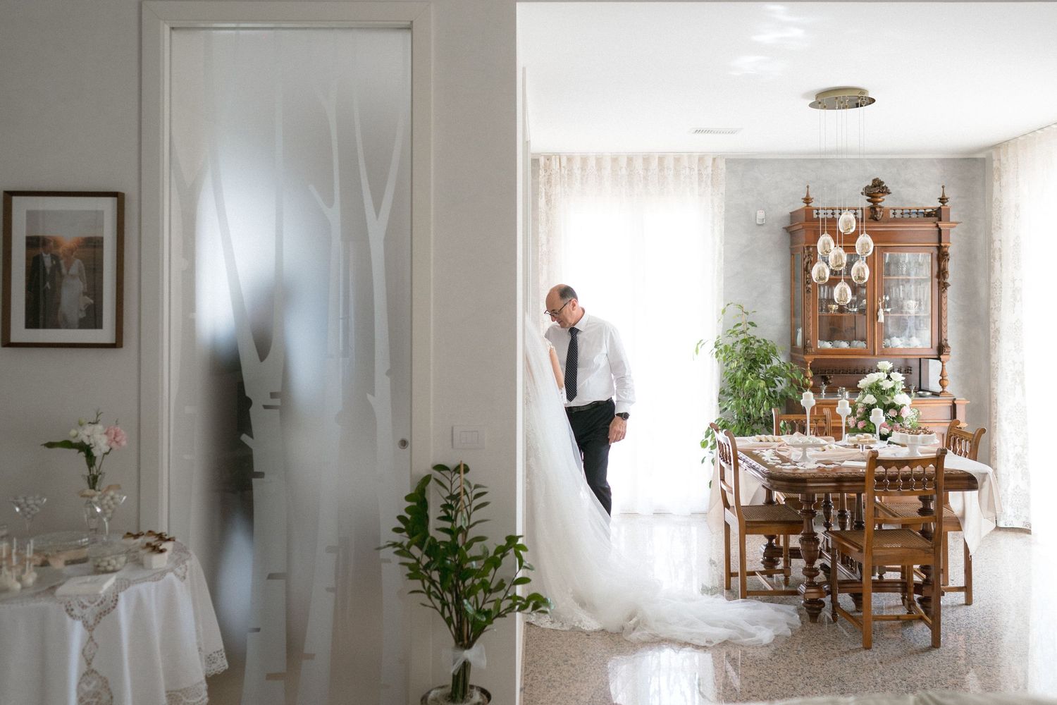 A wedding dress hangs in a bright room with antique furniture and dining table visible through an open doorway.