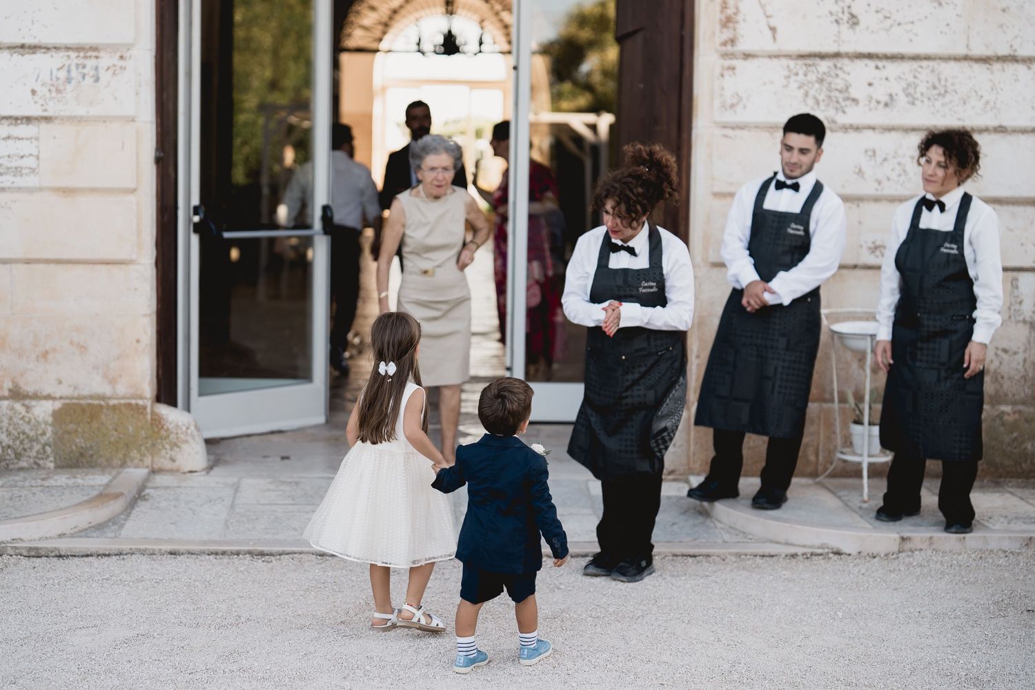 Two small children approach event staff in aprons outside an elegant venue entrance.