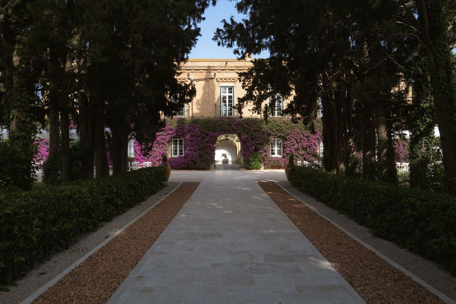 Outdoor wedding venue walkway lined with trees and flowering bushes leading to a stone building.