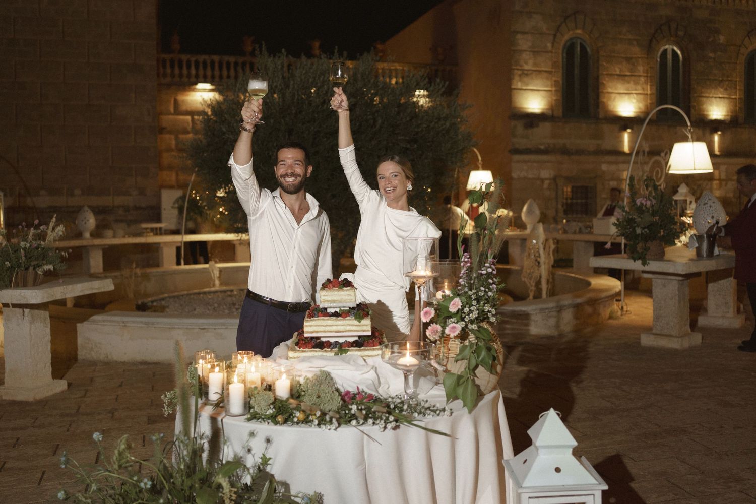 Two people celebrating at an outdoor evening reception with candles and decorative table settings.