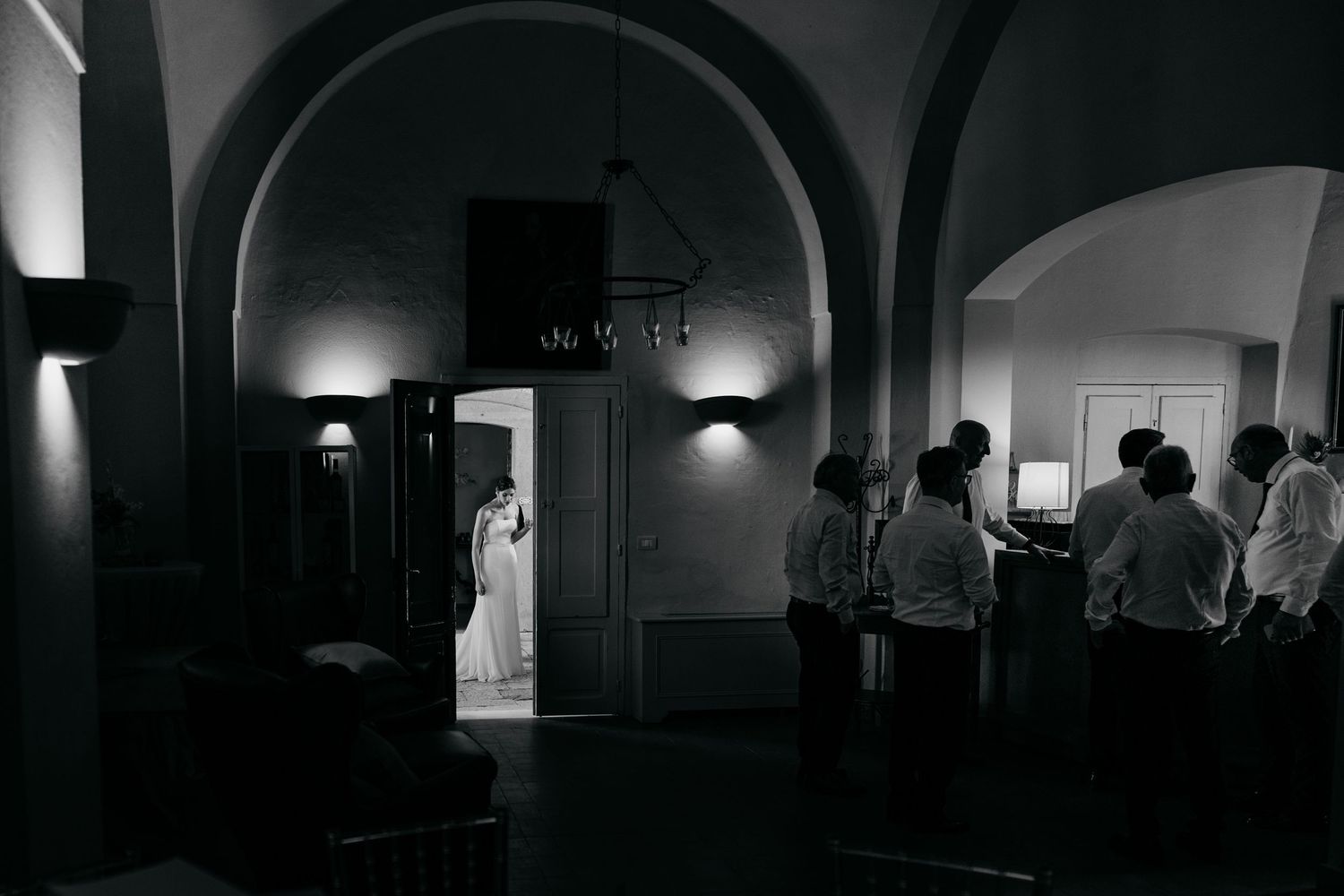 Black and white wedding photo of bride in doorway with groom and groomsmen waiting in dimly lit venue.