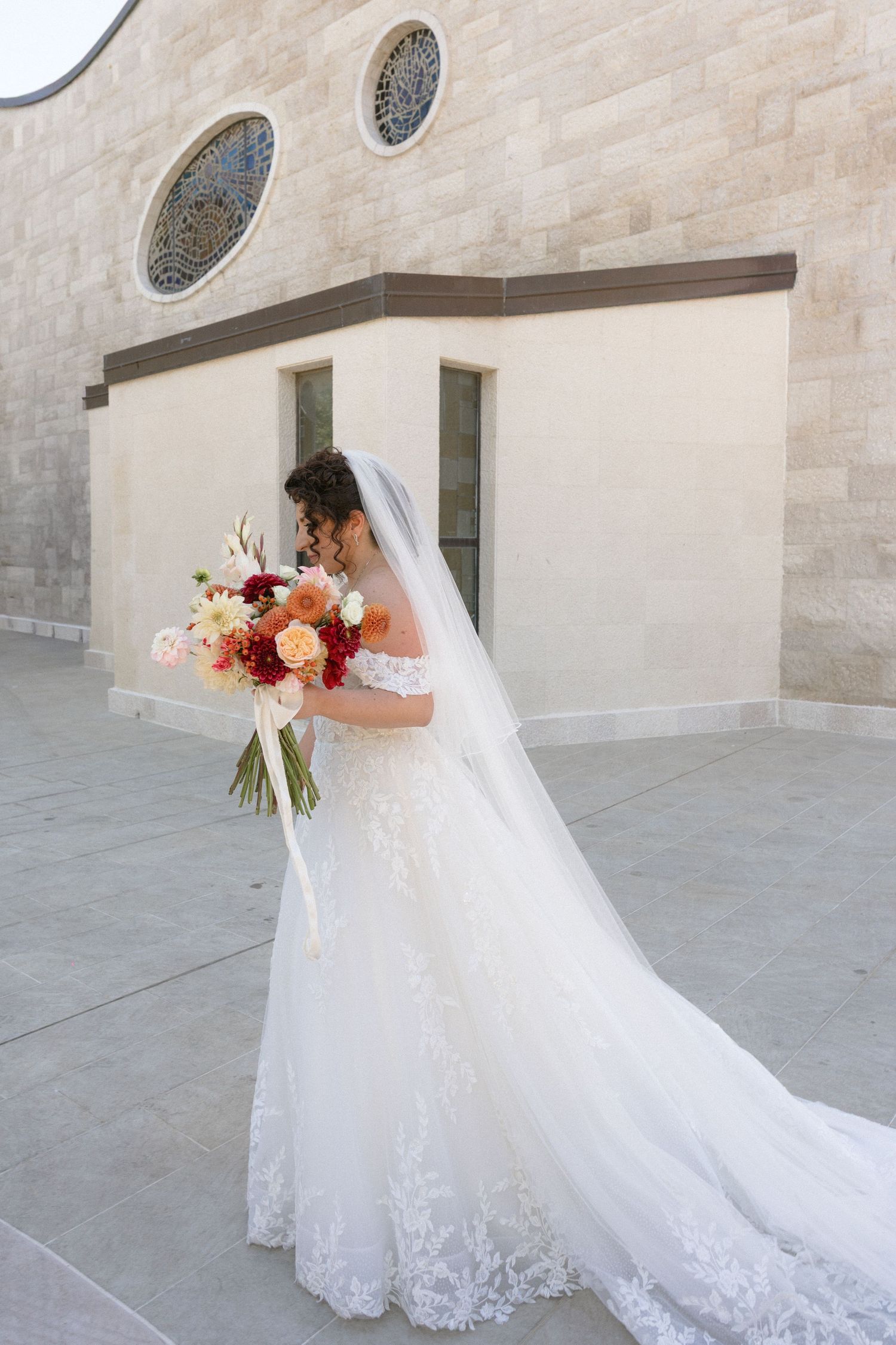 Bride in lace wedding gown with veil holds colorful bouquet of pink and coral flowers outside church.