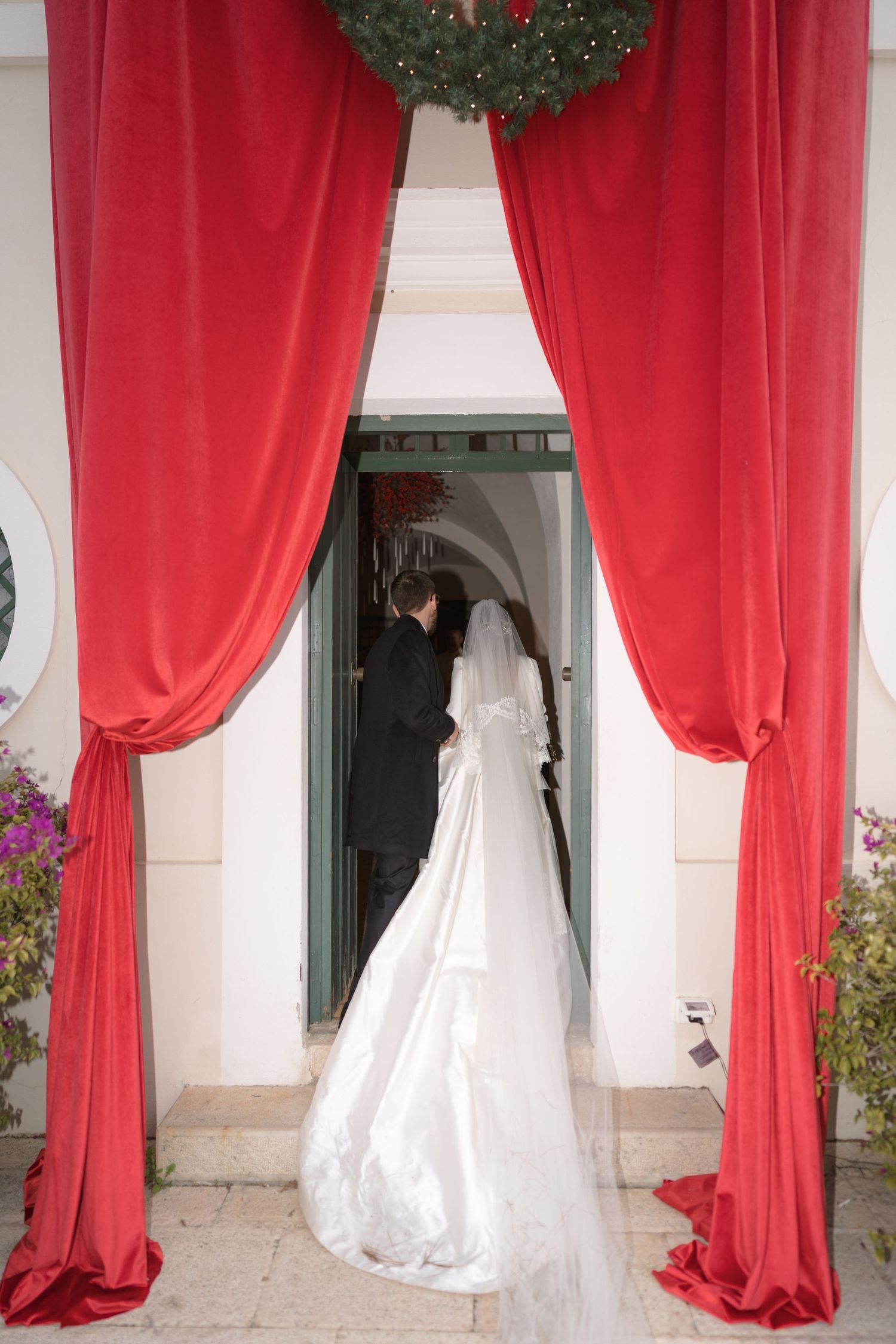 Bride in white gown and groom entering church through red velvet curtains with holiday wreath.