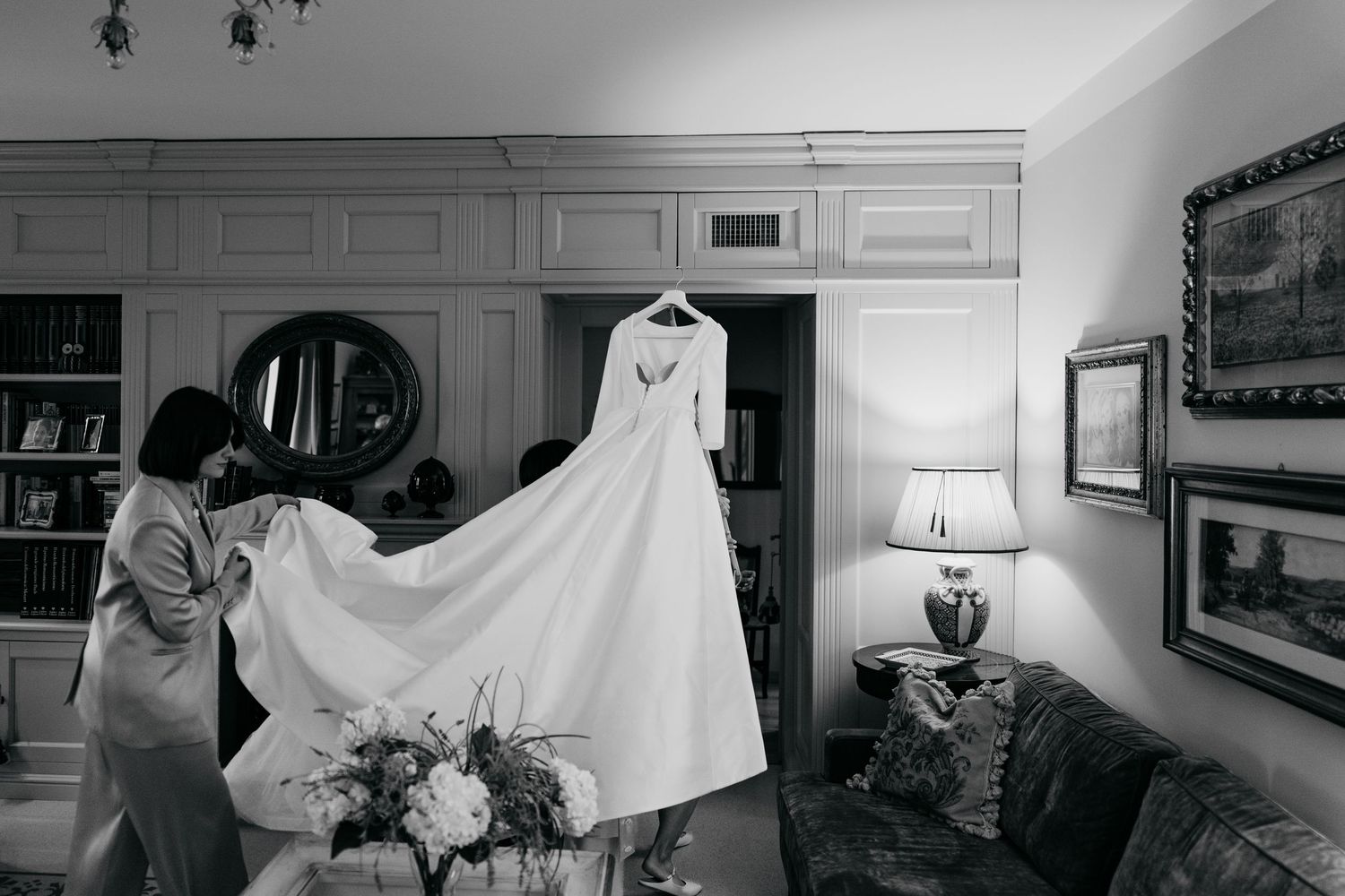 A woman in a robe carefully spreads the train of an elegant white wedding gown in a warmly lit room.