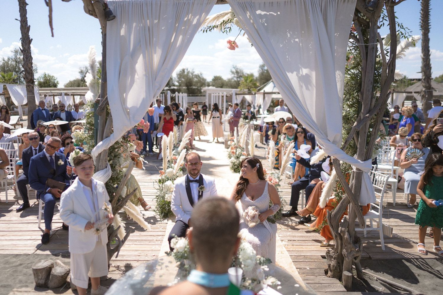 Beach wedding ceremony setup with white fabric draped archway and palm trees.