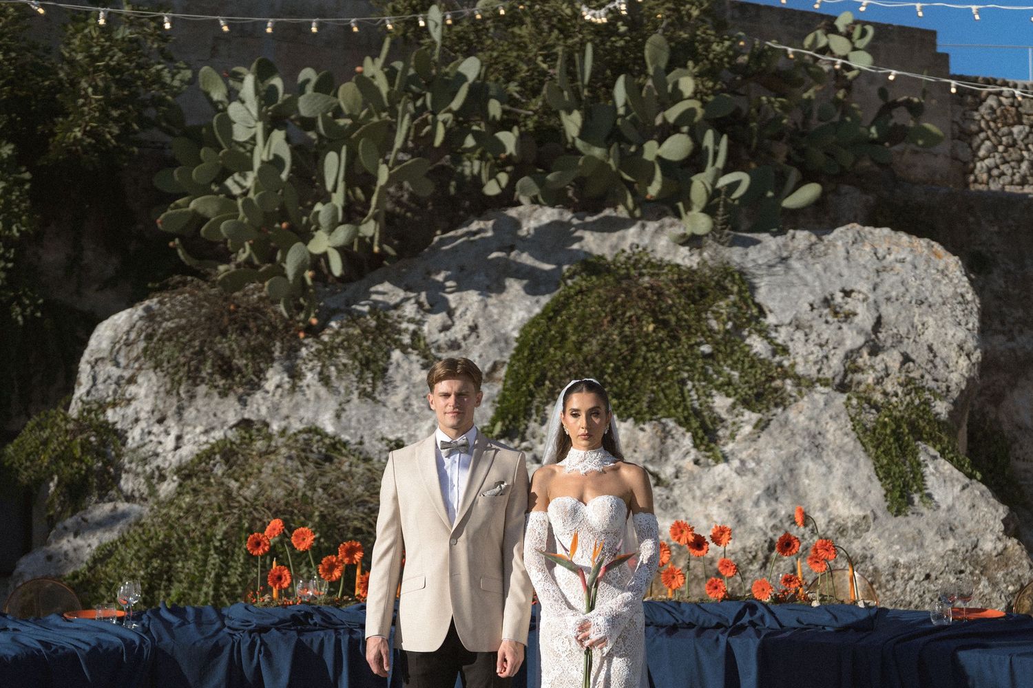 Bride and groom in a beige blazer stand together before a large rock formation with orange flowers on a blue-draped table.