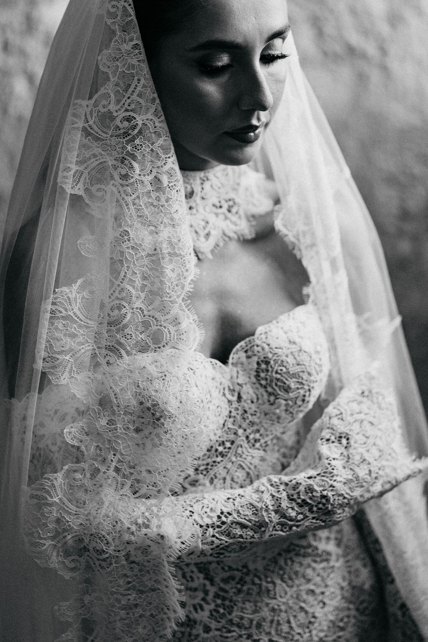Black and white close-up of a bride gazing downward in a lace wedding dress with a long lace-trimmed veil.