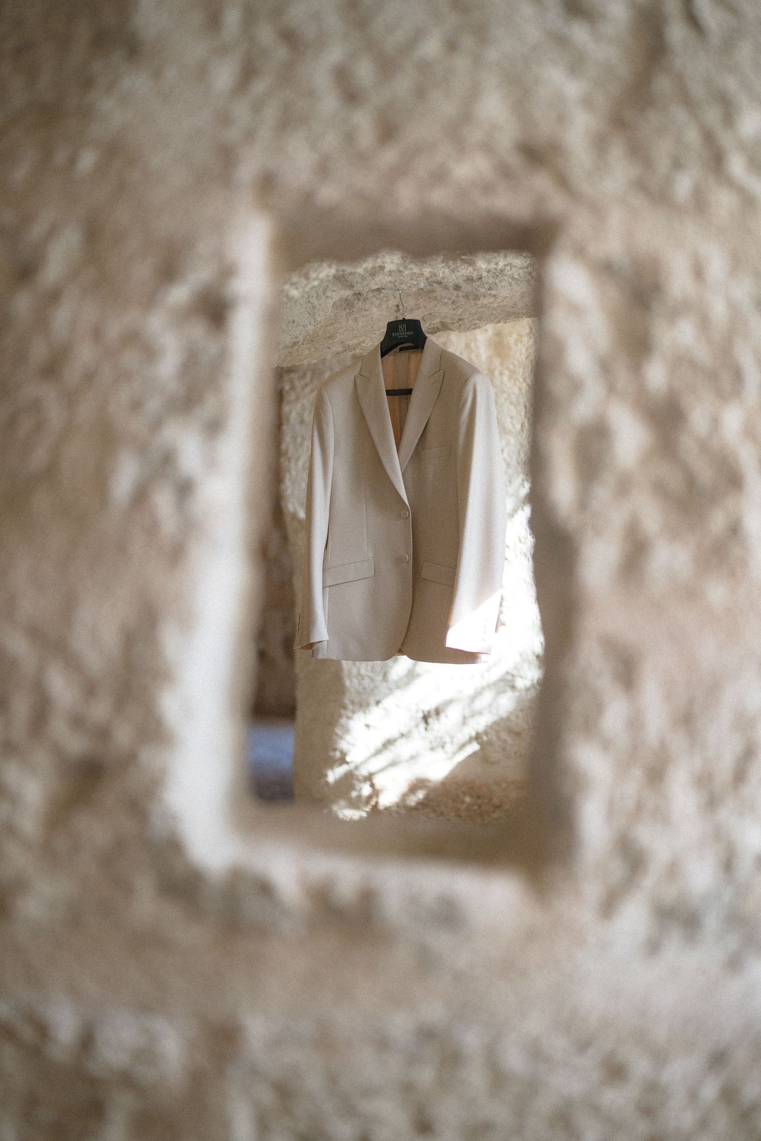 Cream wedding suit jacket hanging on a hanger, framed through a window opening in a rustic stone wall.