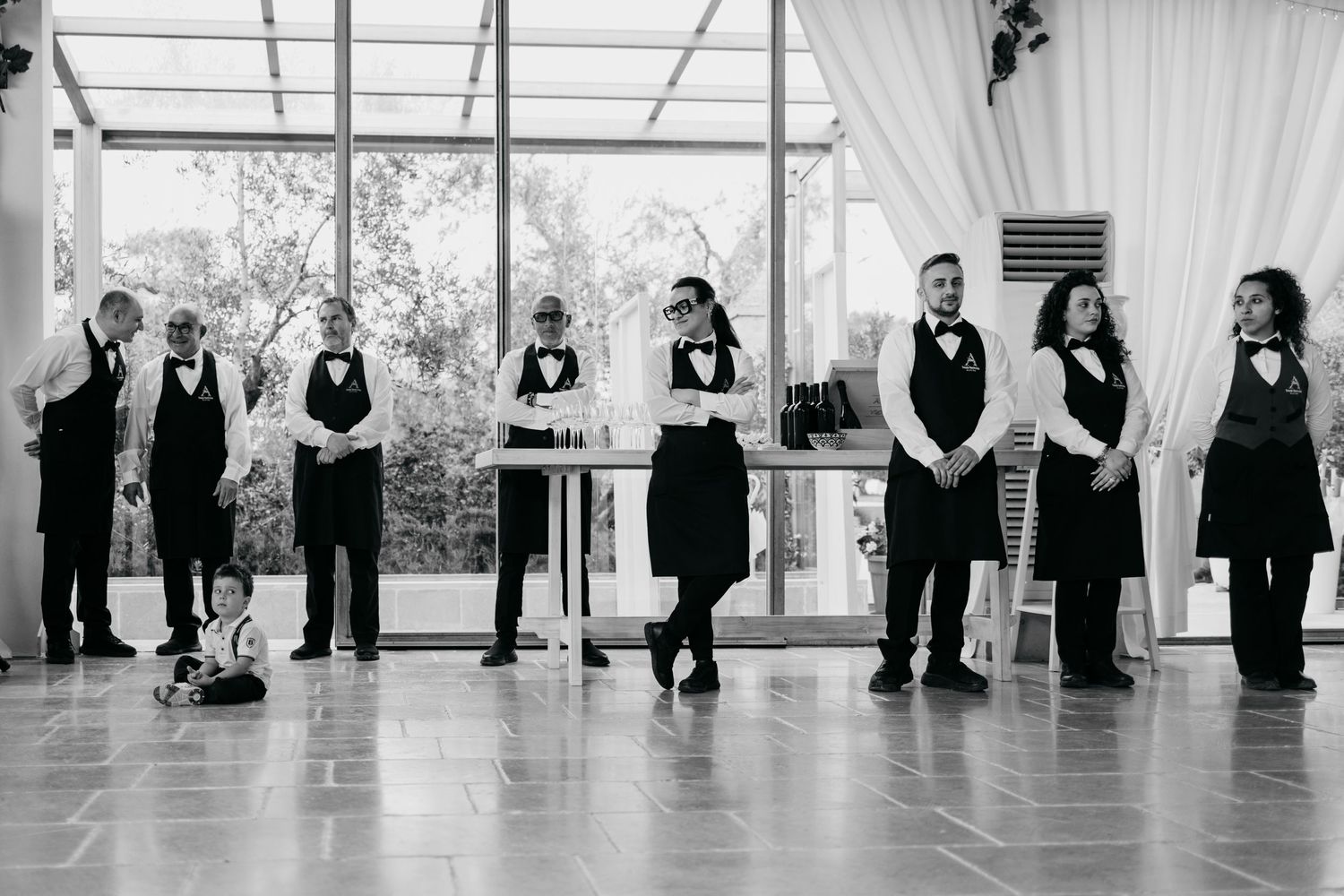 Wedding party members lined up in black attire against bright windows for a formal photo.
