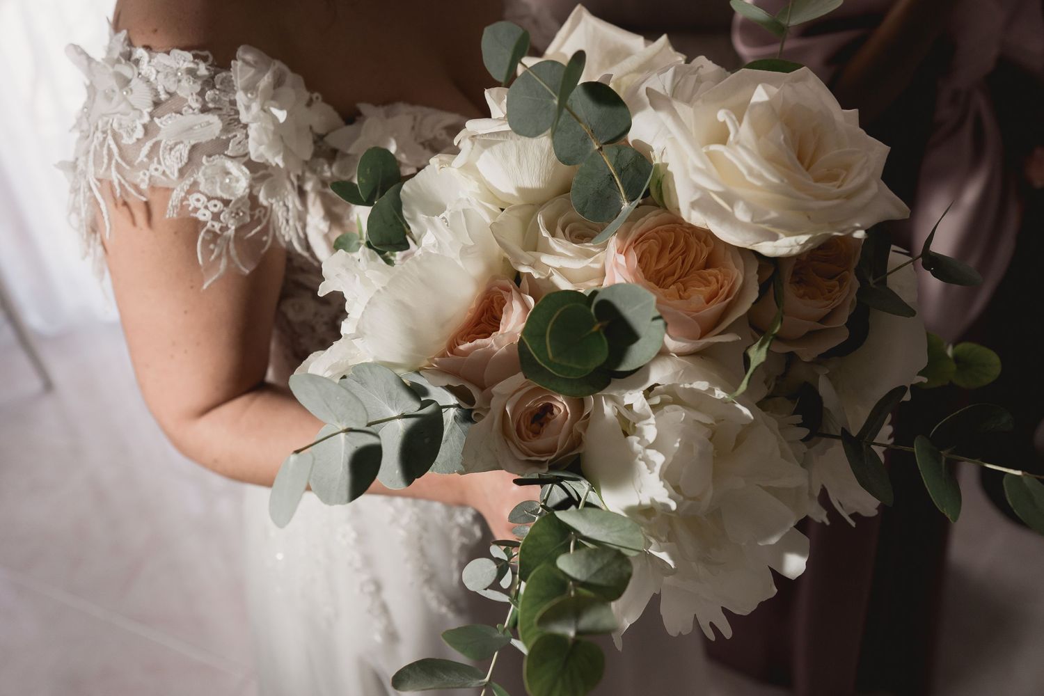 Close-up of an elegant white and blush wedding bouquet with eucalyptus held by someone in a lace dress.