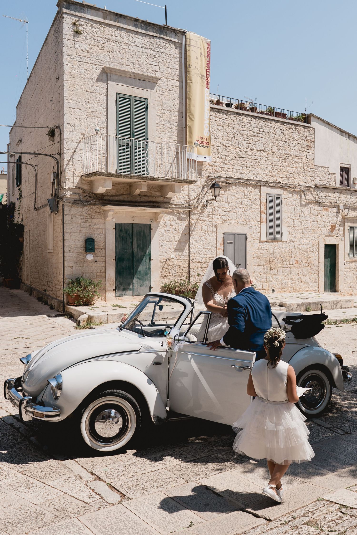 Wedding couple posing with a vintage white Volkswagen Beetle against Mediterranean architecture.