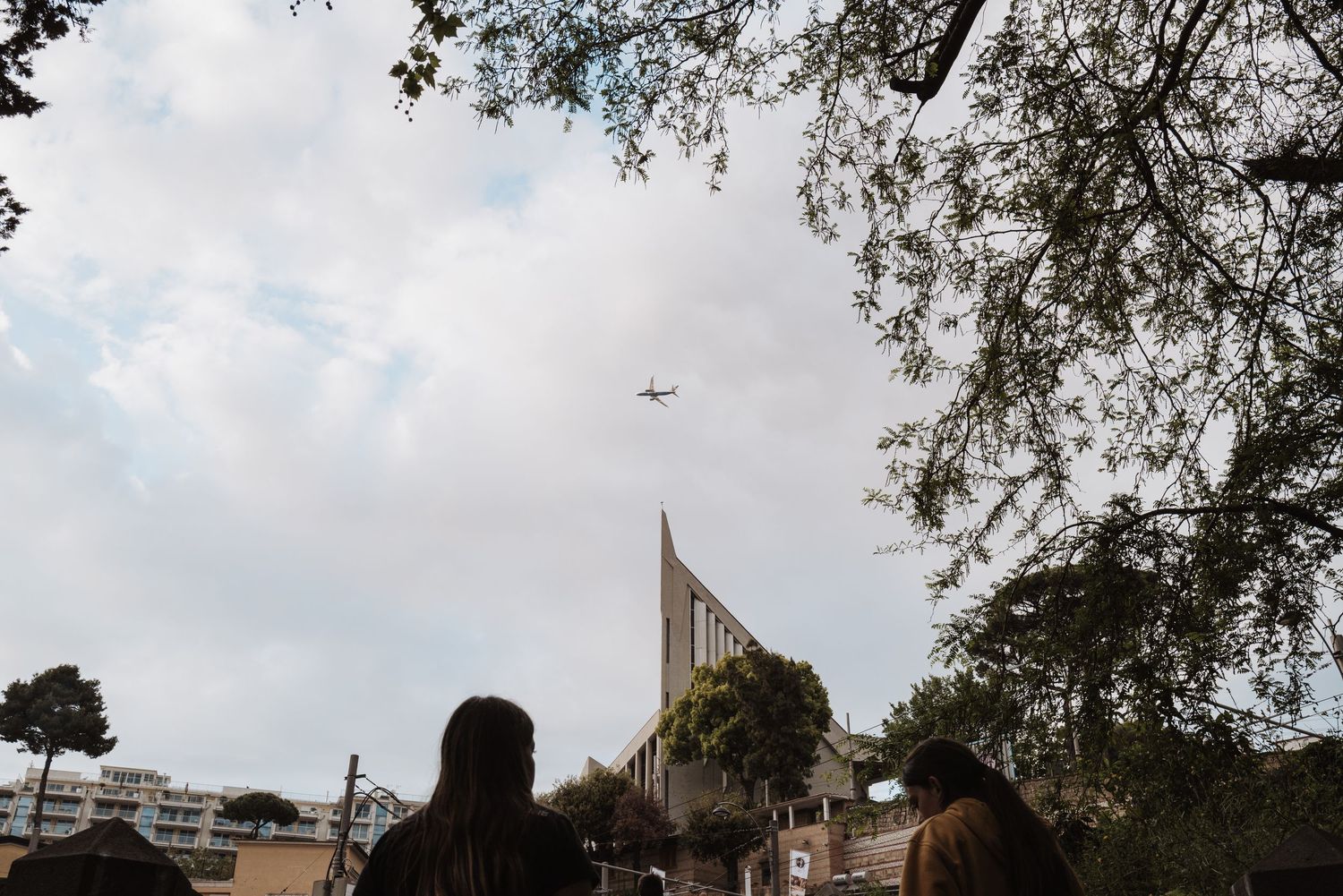 People gather beneath trees with a modern architectural structure and airplane visible in the cloudy sky.