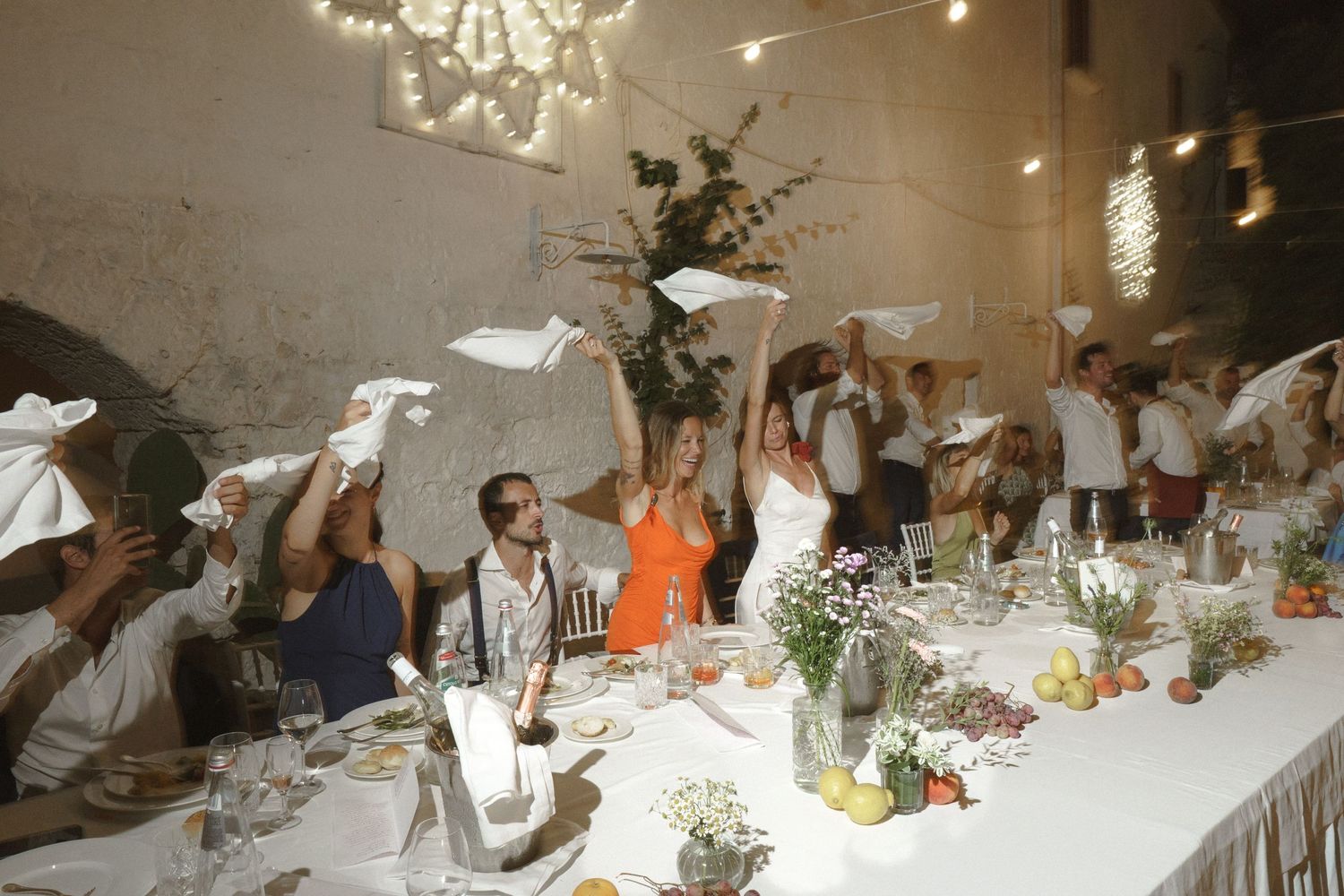Wedding reception guests wave white napkins in celebration at a long decorated dining table with floral centerpieces.