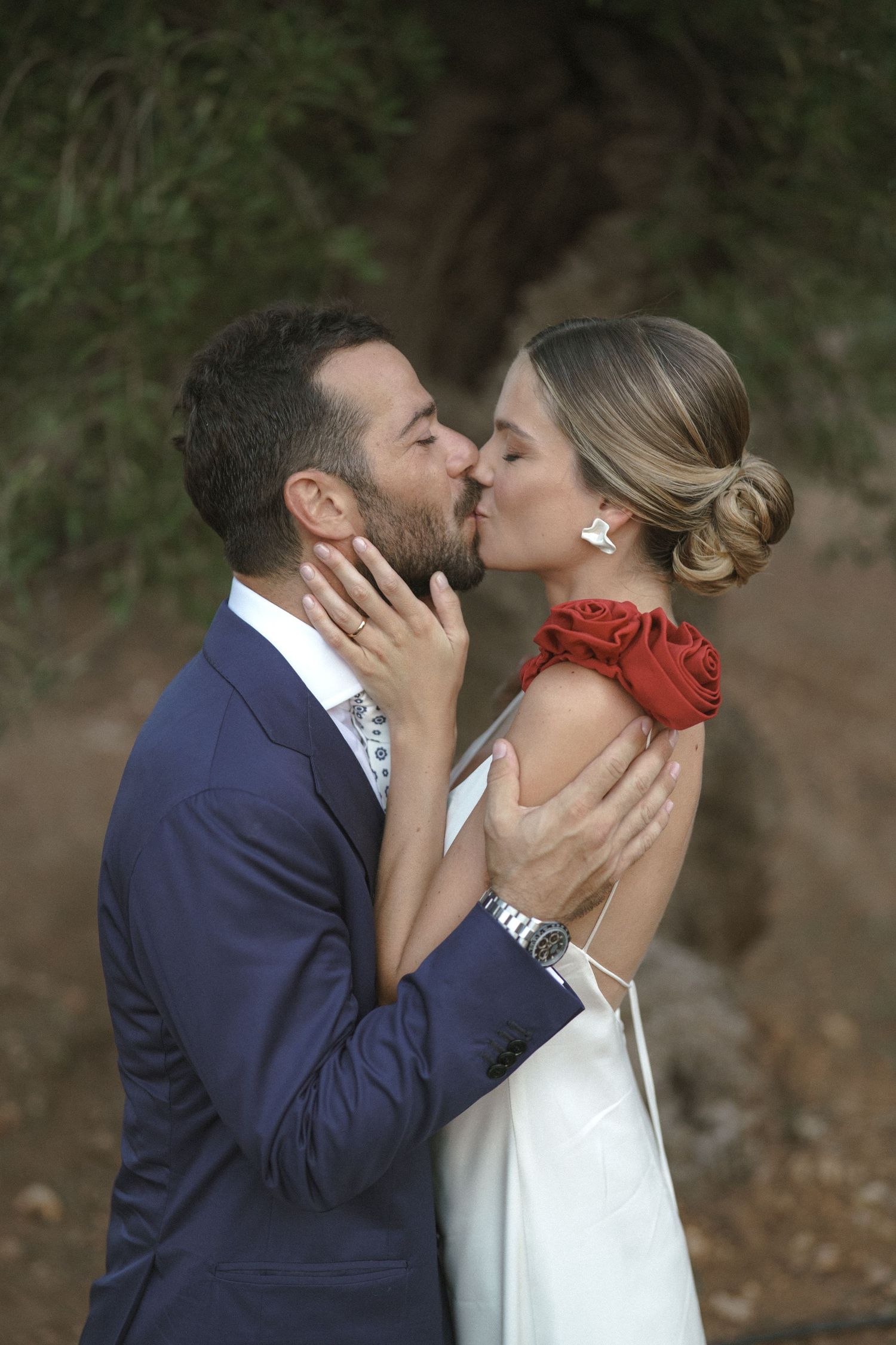 Romantic wedding portrait with a couple kissing, bride wearing elegant white dress and red rose corsage, groom in navy suit.