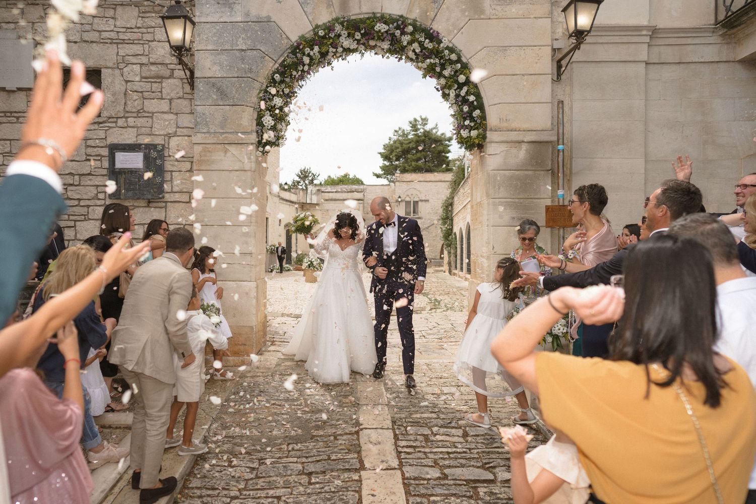 Guests throw petals as newlyweds exit under a floral arch at an outdoor stone venue.