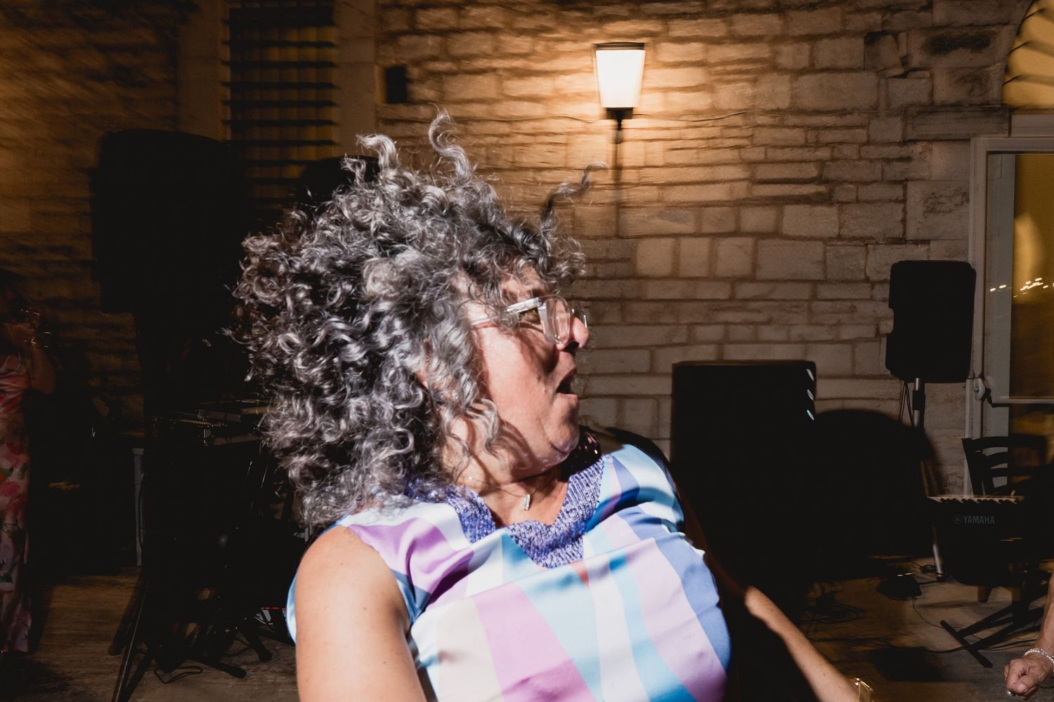 Person with curly gray hair wearing a tie-dye shirt laughs joyfully against a brick wall with dim lighting.