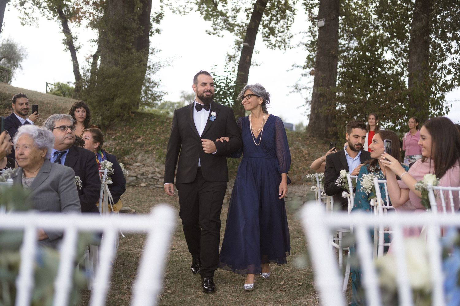Wedding guests gather outdoors near white chairs during an outdoor ceremony.