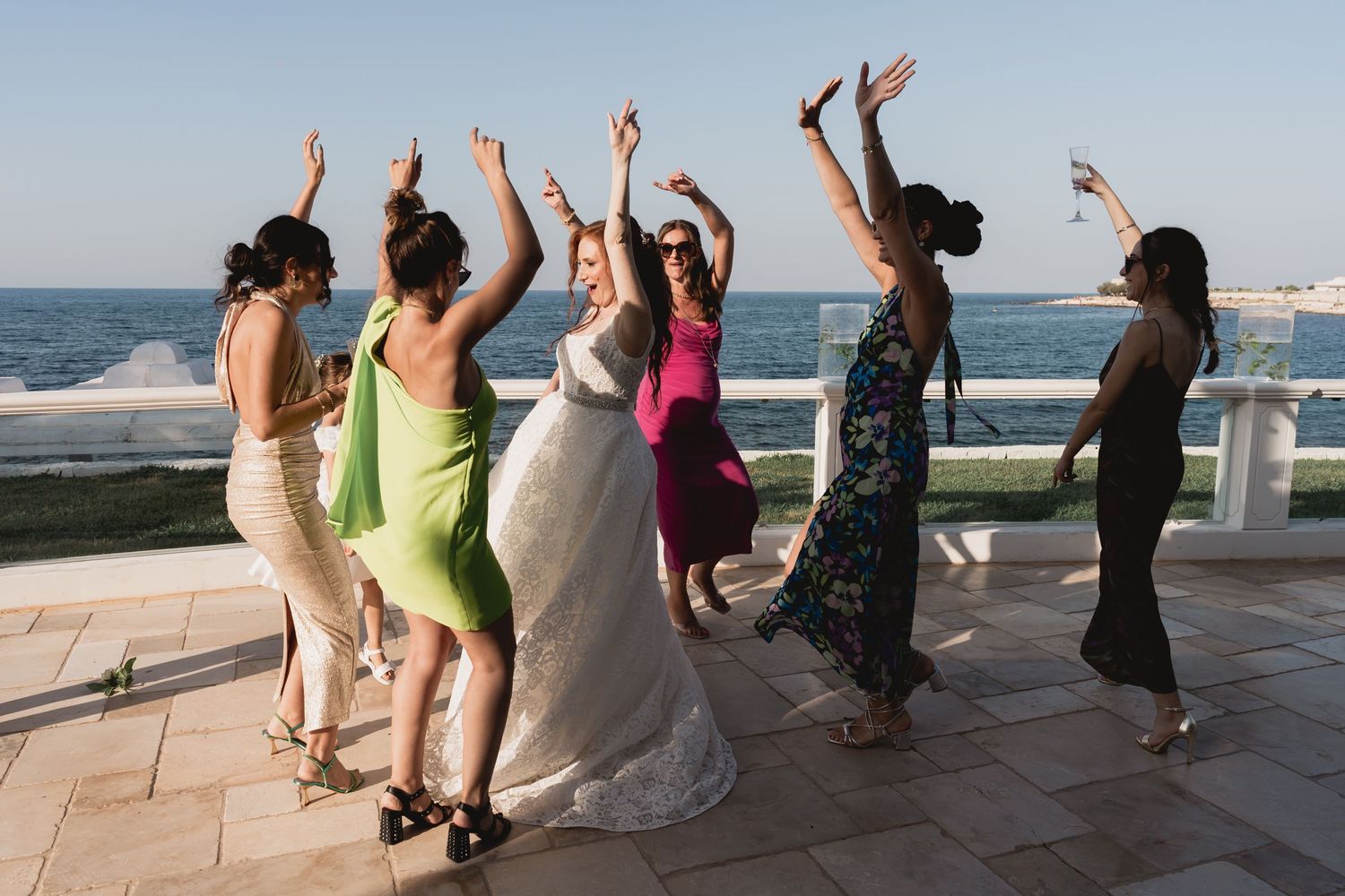 Wedding party dances joyfully on a seaside terrace during sunset celebrations.