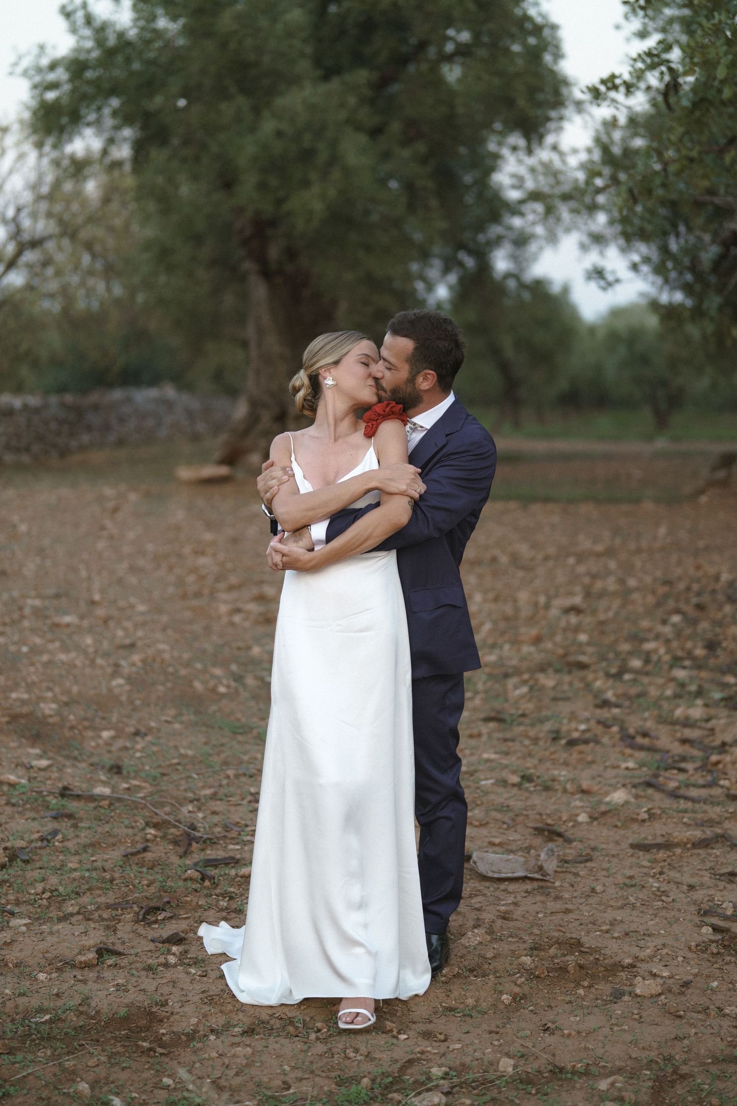 A romantic wedding moment captured with a couple embracing on rustic ground surrounded by olive trees.