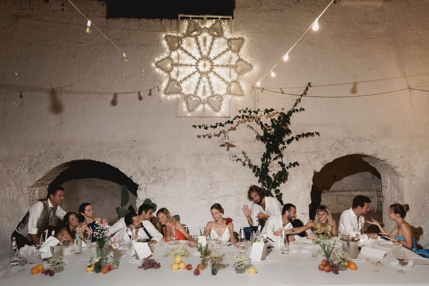 Wedding reception dinner under string lights with guests seated at long tables beneath ornate ceiling decoration.