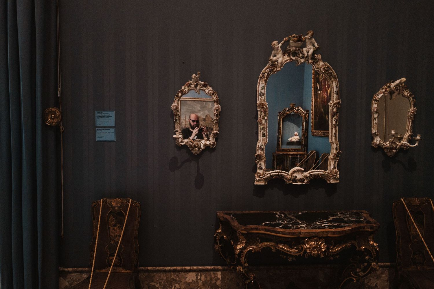 Ornate vintage gold mirrors and console table against a dark wall with teal curtains in a classic interior setting.